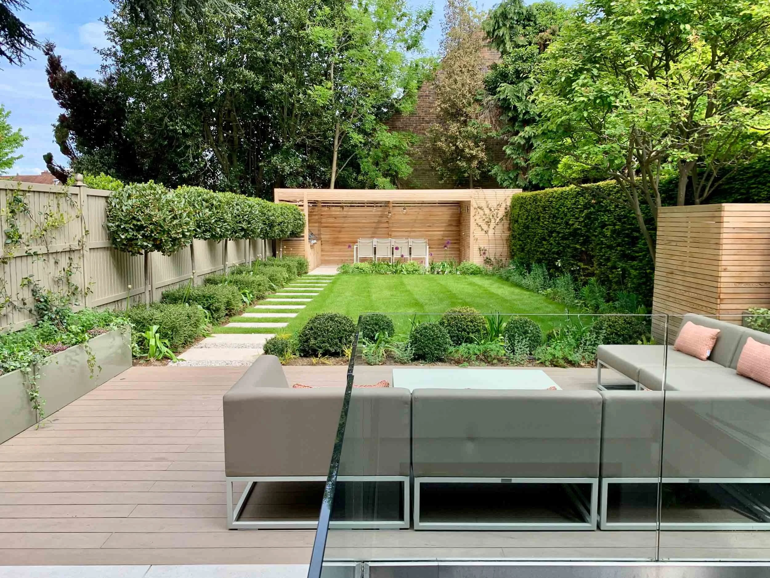 Modern backyard with wooden deck, glass railing, outdoor seating, manicured lawn, stepping stones, garden beds, and tall trees.