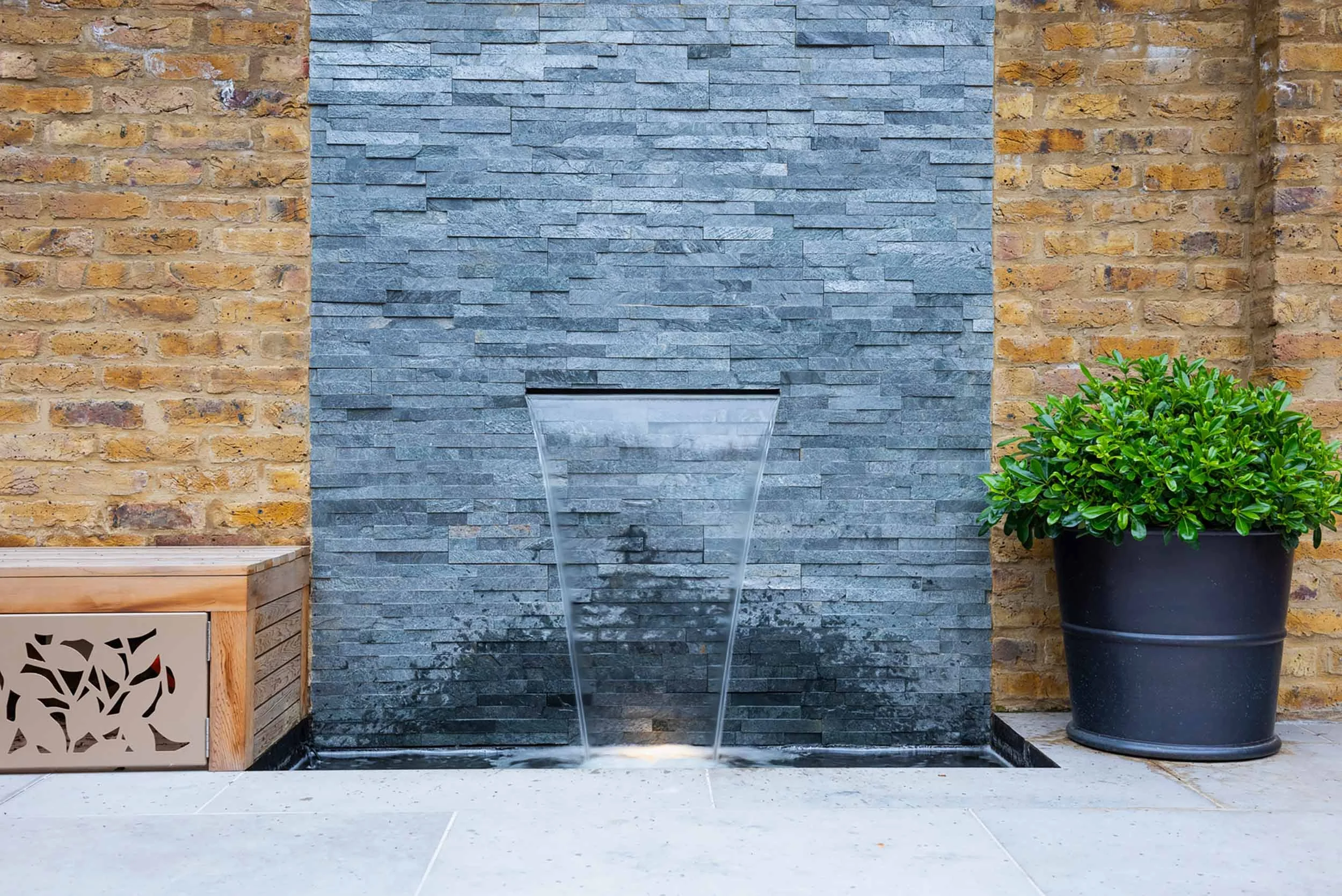 Modern outdoor water feature with water flowing from a tall, narrow, curved glass structure onto a black basin, flanked by a brick wall and a potted green shrub.