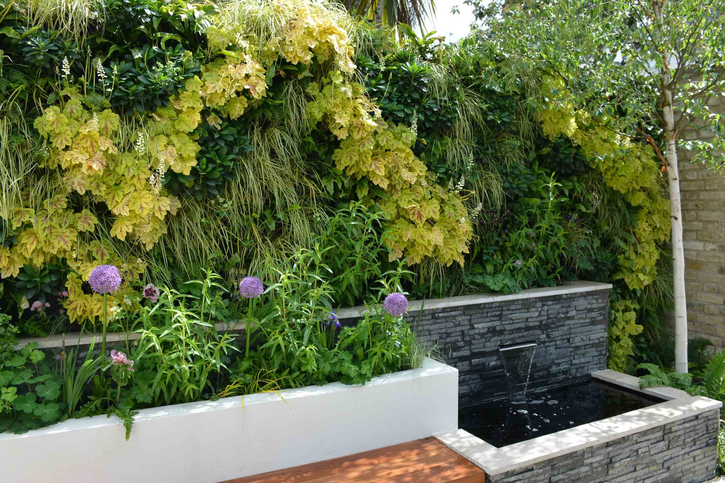 A lush green garden with a variety of plants, including yellow-green foliage, purple flowers, and a small water fountain in a black stone planter, against a brick wall on the right.