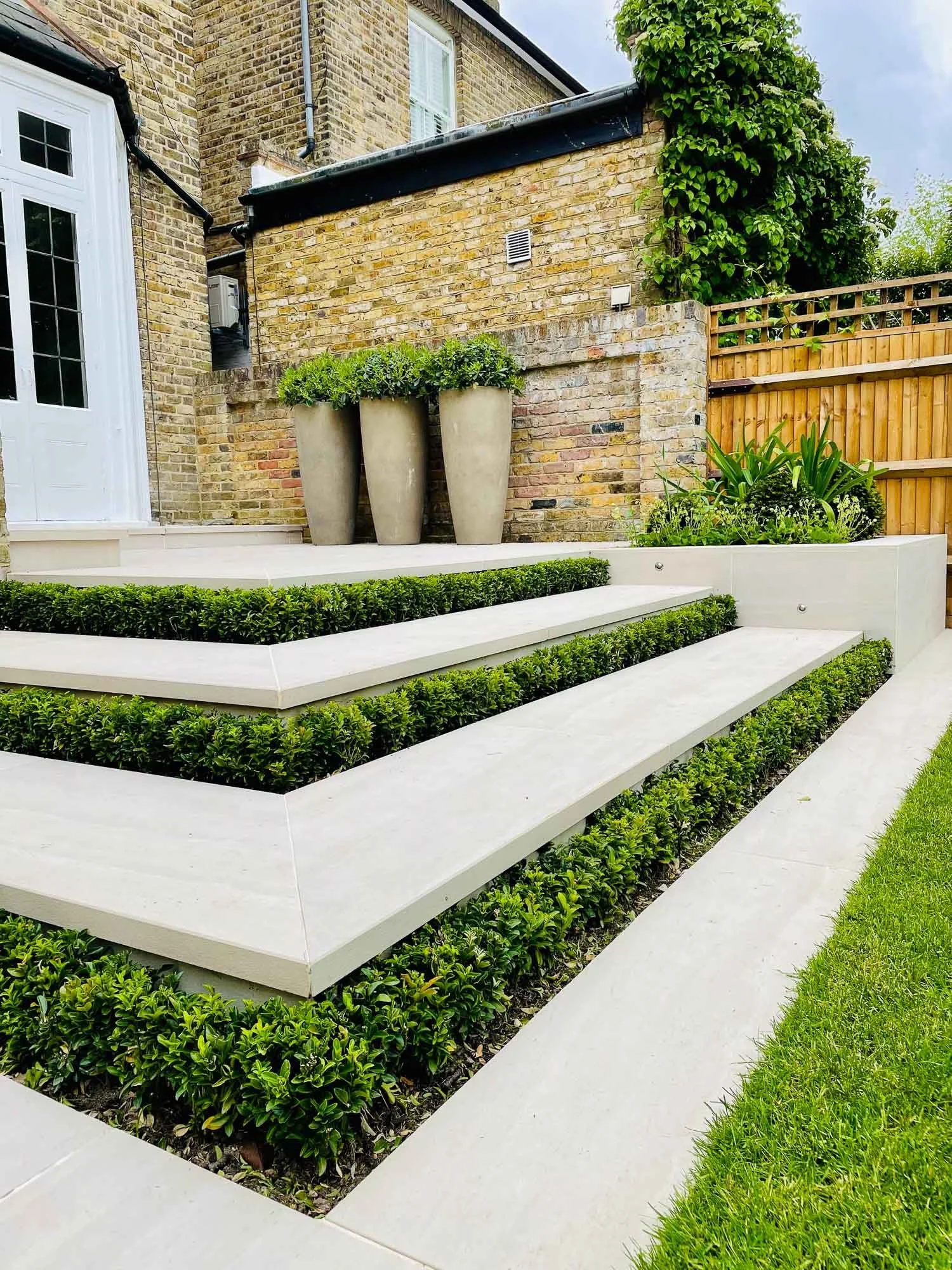 Modern garden with tiered white concrete steps, green shrubbery, large beige planters with greenery, and a brick wall with an ivy plant.
