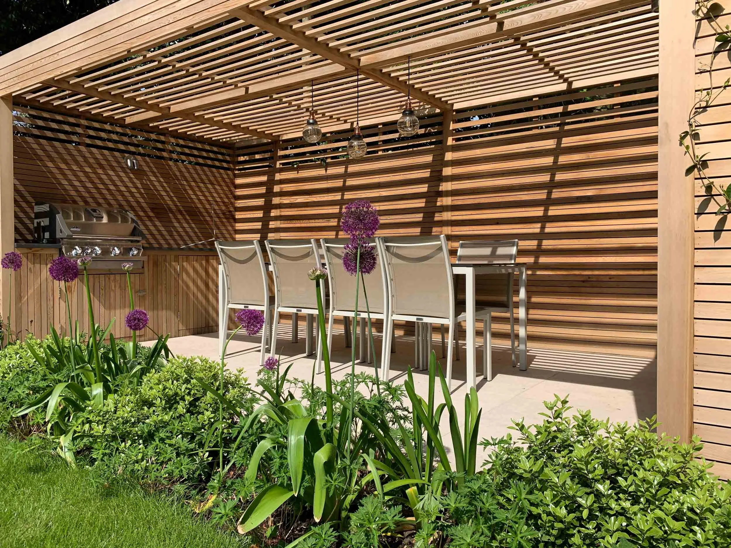 Outdoor patio with a dining table and six chairs under a wooden pergola, surrounded by plants with purple flowers and green grass.