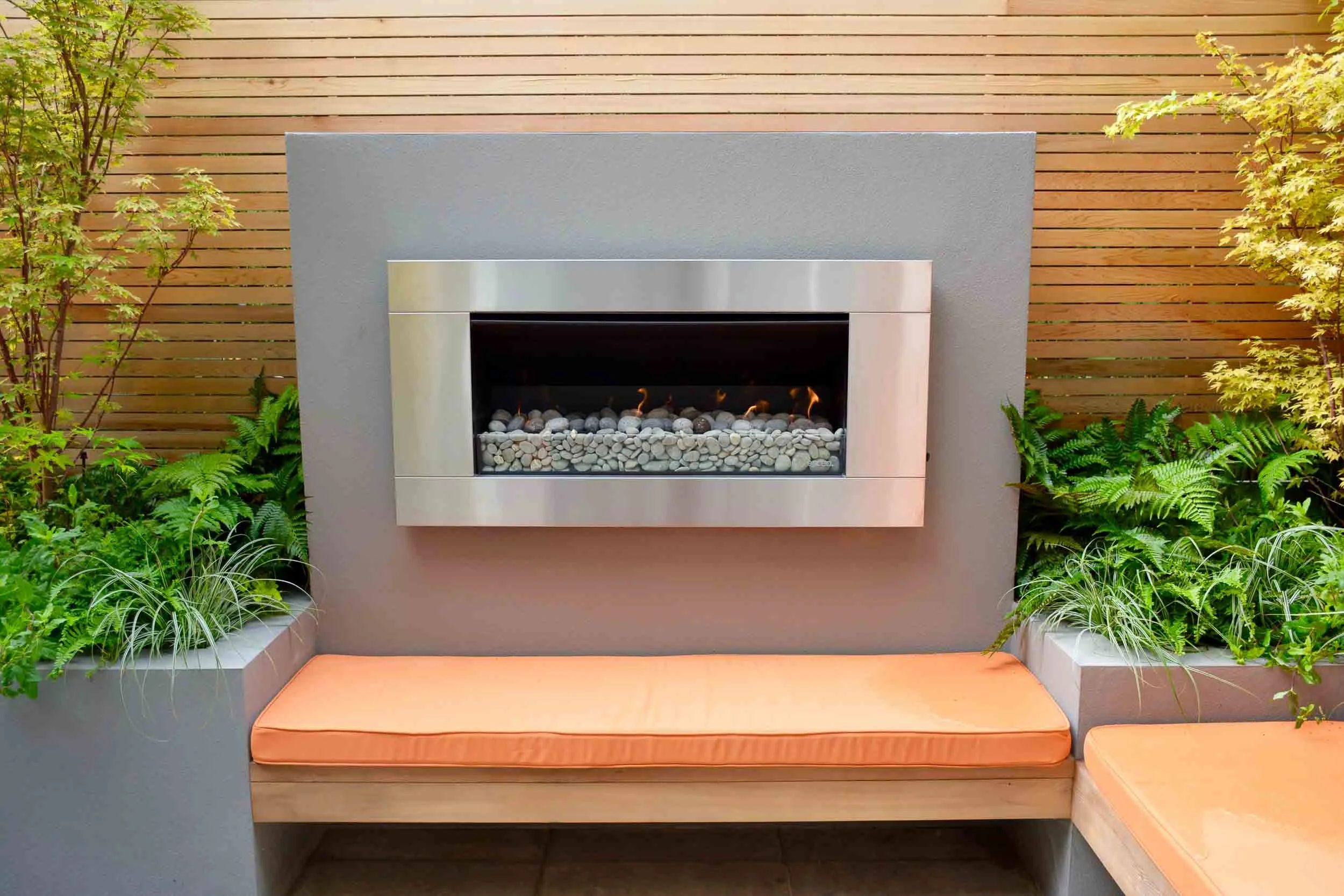 Modern outdoor fireplace with orange cushioned seating, surrounded by green plants and a wooden fence.