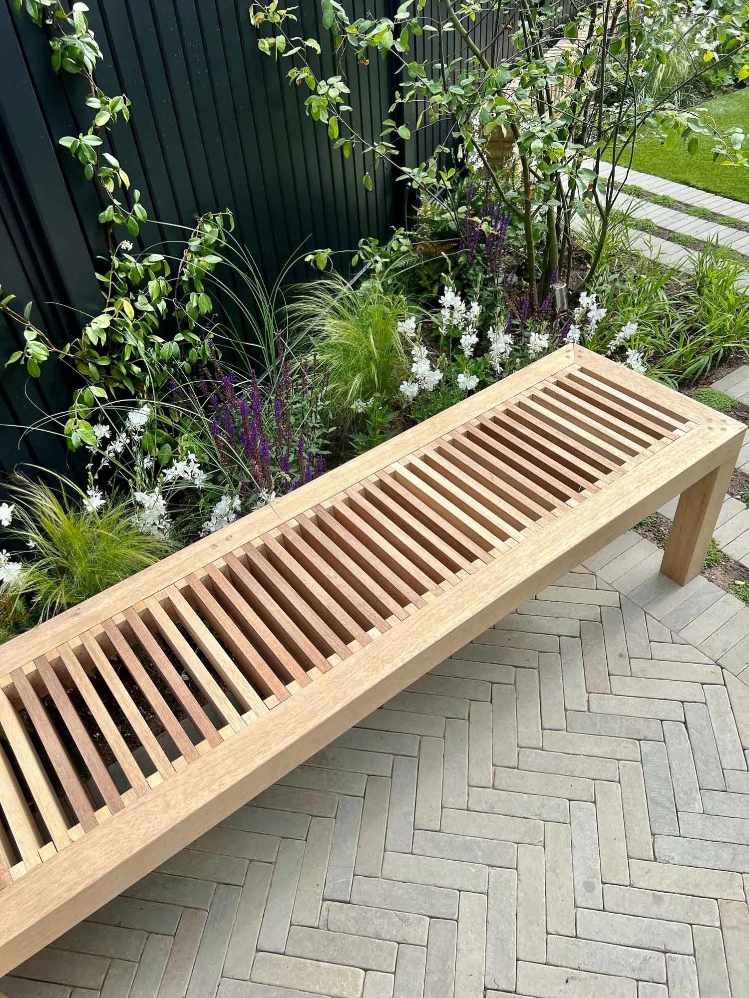 Wooden slatted garden bench on paved patio, next to a flower bed with white and purple flowers and green foliage, against a dark fence and grassy lawn.