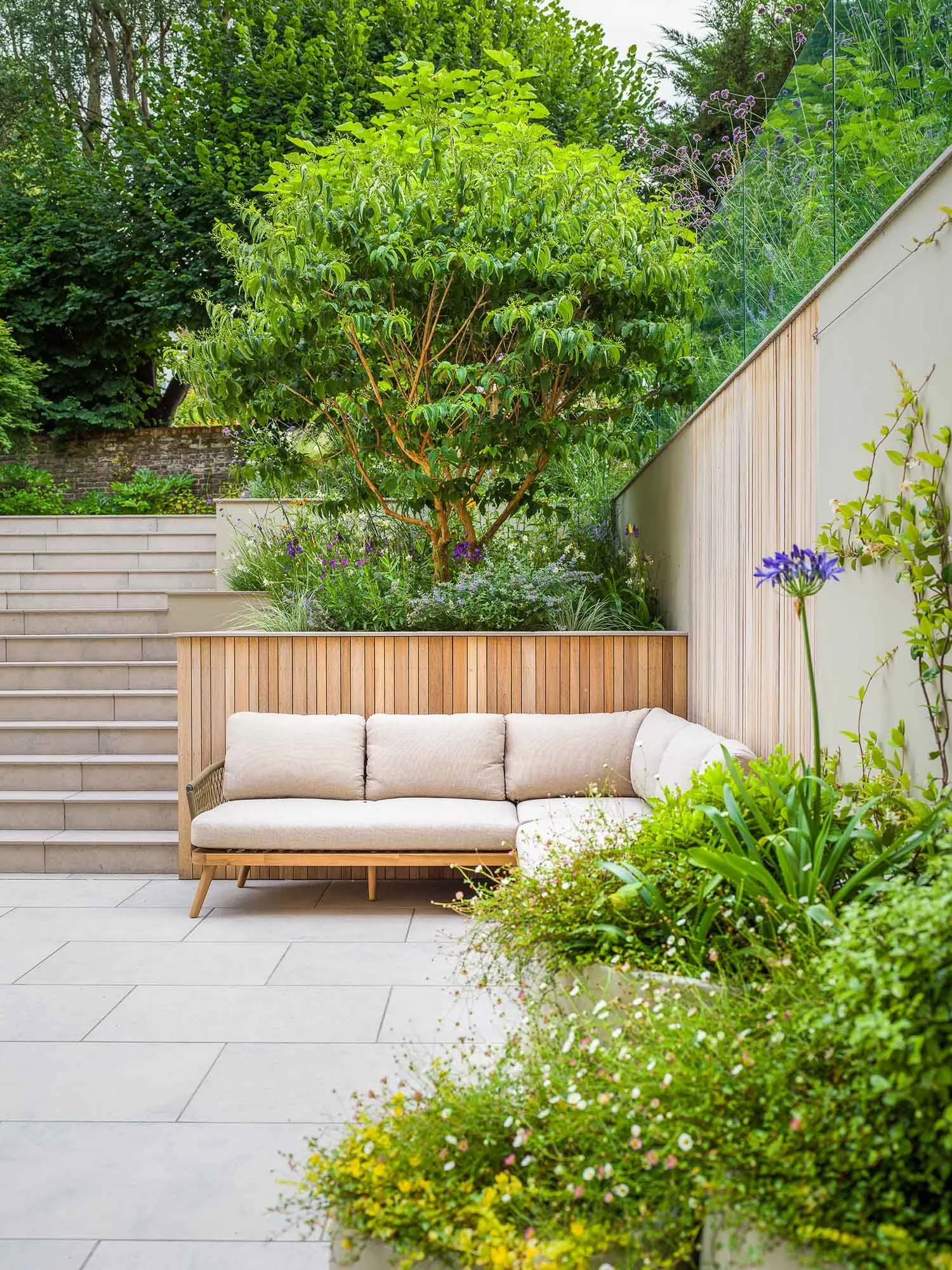 Modern garden terrace with a cream L-shaped sofa and light stone paving next to a wide staircase and lush green planters