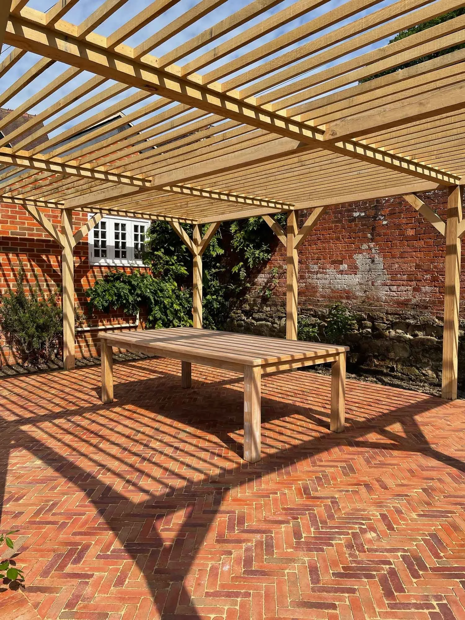 Unfinished wooden pergola over a brick patio with a picnic table and some plants in the background. Shadows from the pergola create pattern on the ground.