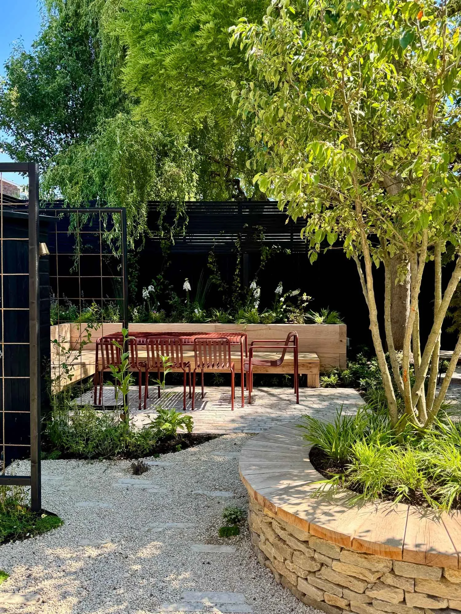 A backyard patio with red chairs, wooden bench, gravel and stone pathway, lush green trees, plants, and shrubs, with a black fence in the background.