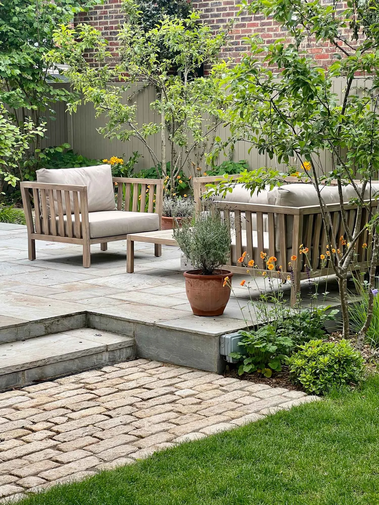 A cozy backyard patio with two beige cushioned wooden chairs and a matching coffee table, surrounded by lush green plants, trees, and a brick wall in the background.