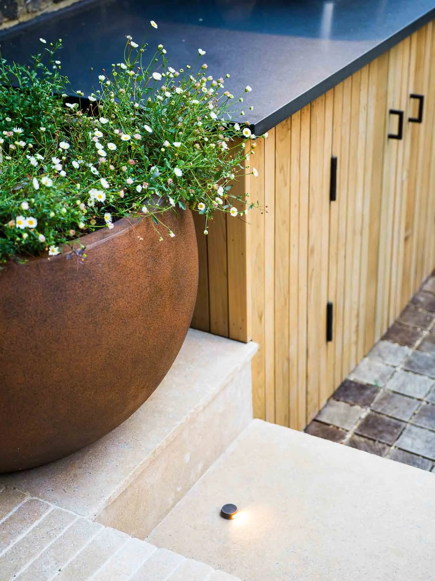 Detail of a small round garden light set into a stone step beside a large corten steel planter