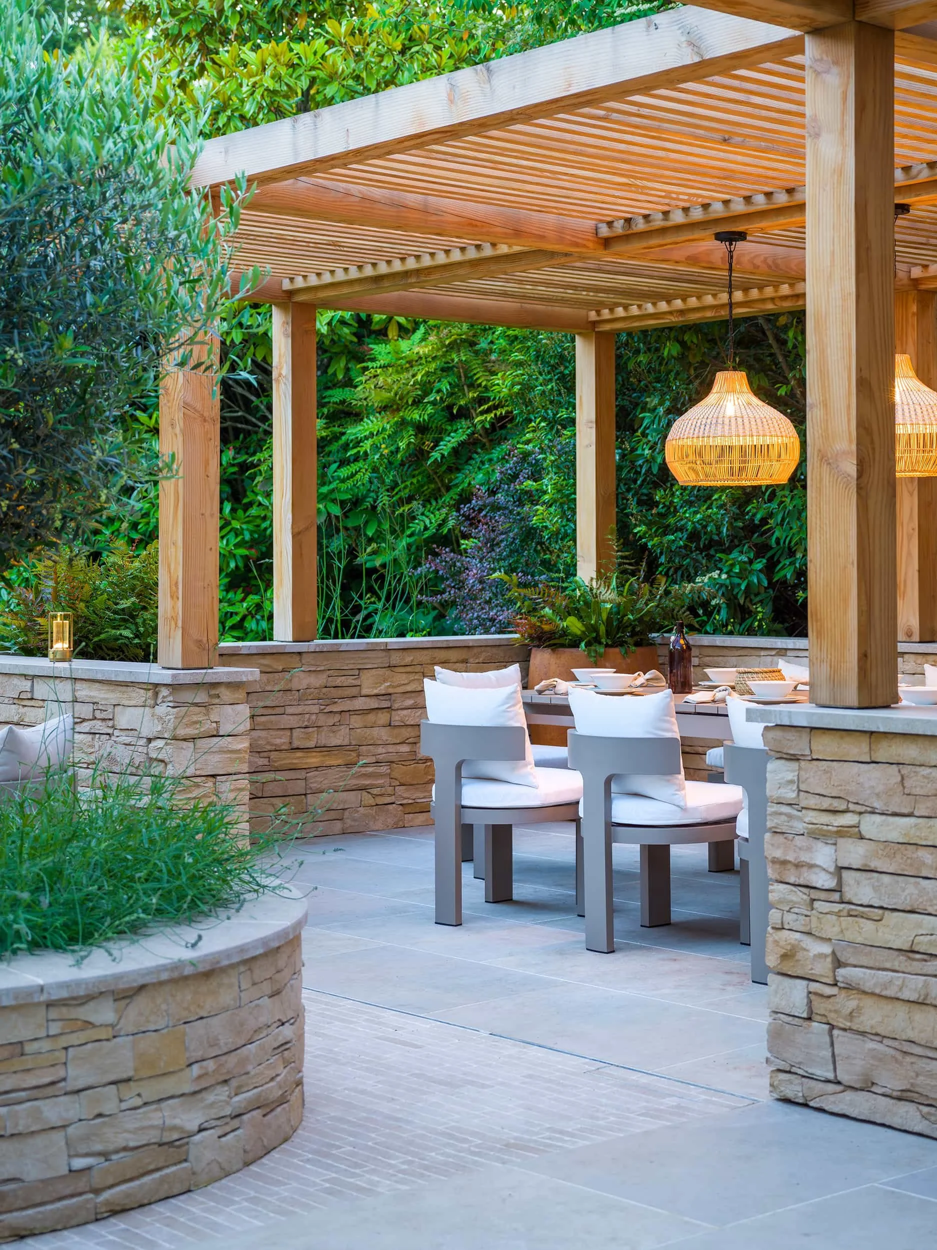 uxury outdoor living space with stone walls and a timber pergola over a dining set with white cushions and lamps