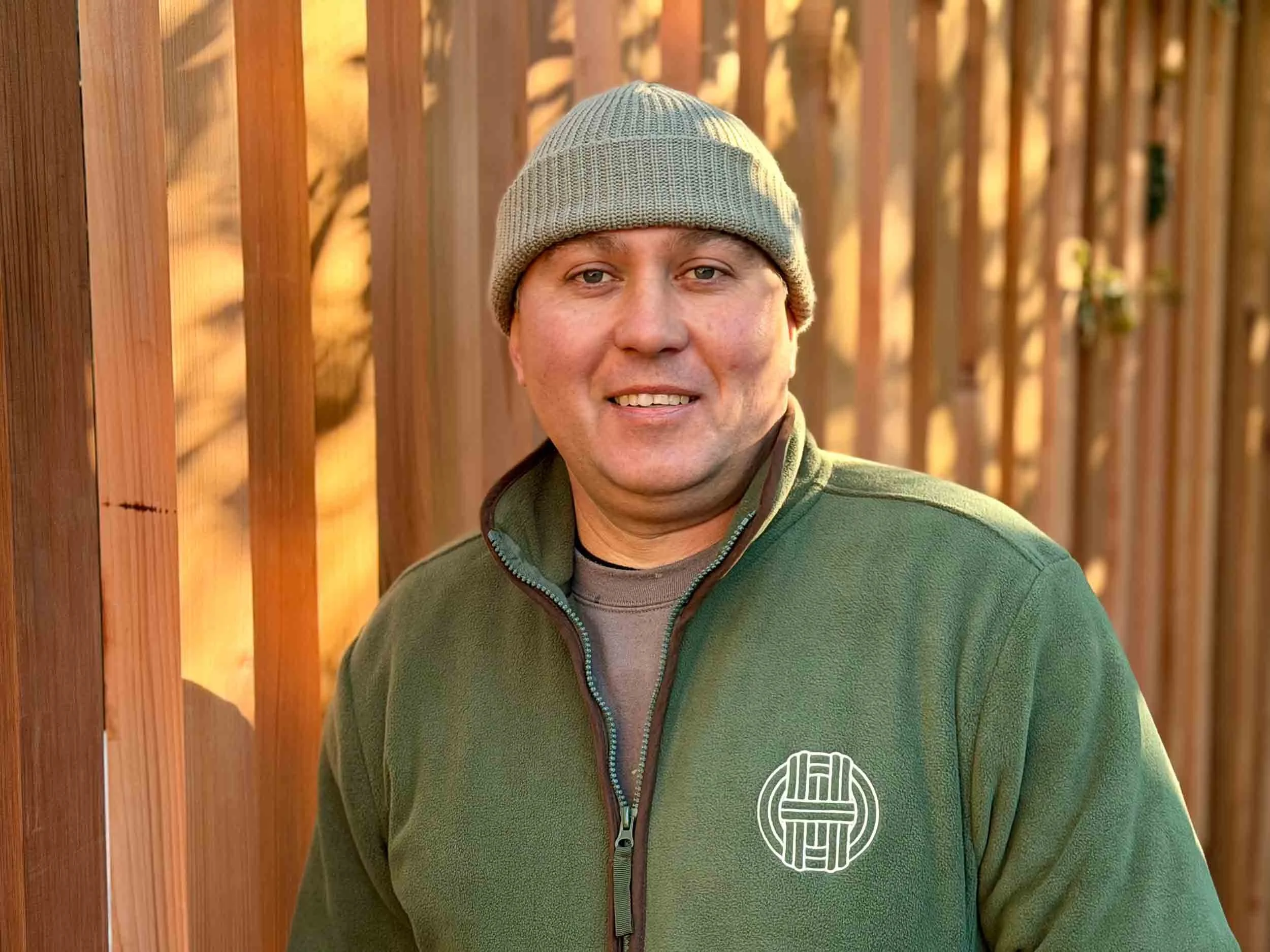 Smiling man wearing a light grey beanie and a green zip-up fleece with a white logo in front of a wooden slat fence