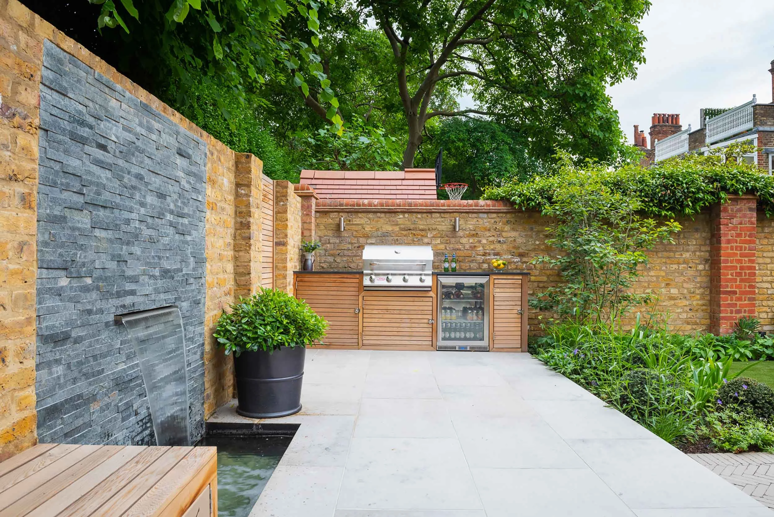 Modern garden with slate water feature stone paving and outdoor kitchen with a stainless steel barbecue and drinks fridge