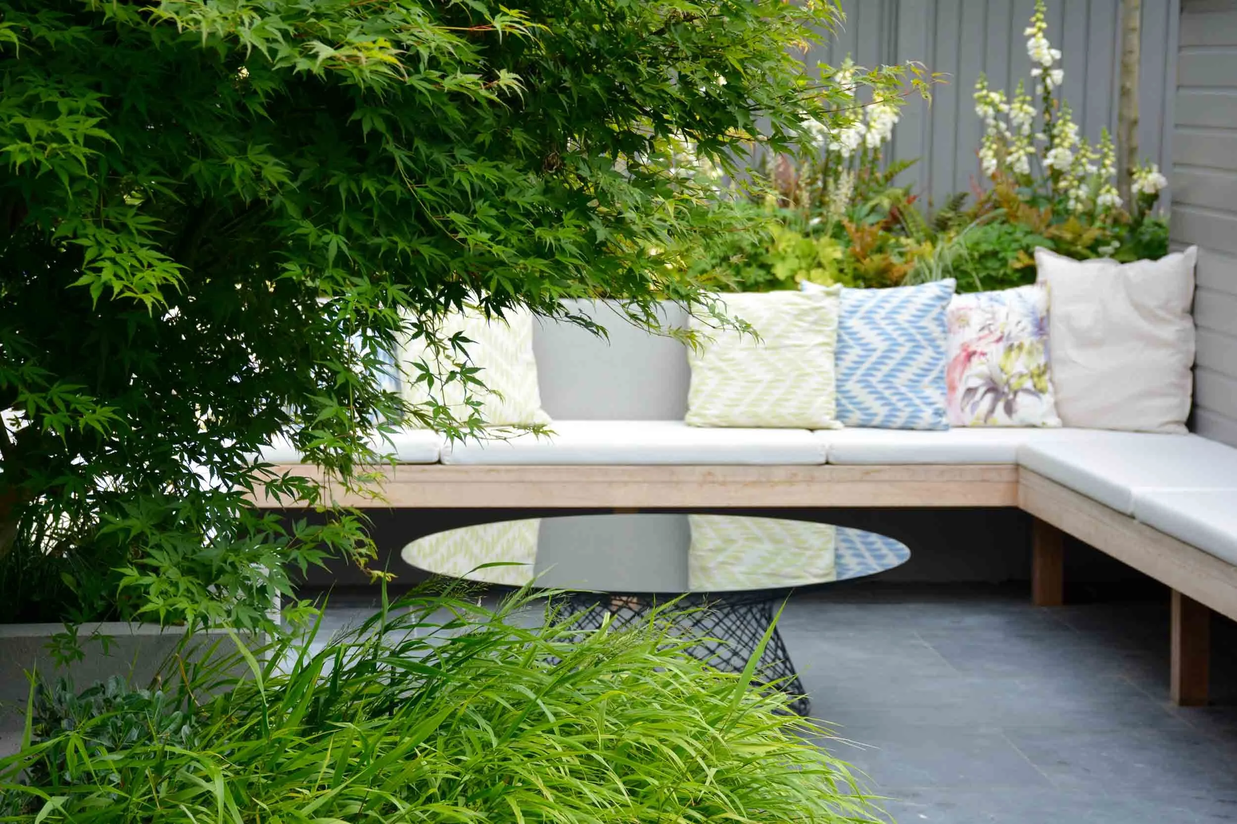 Outdoor patio seating area with a sectional sofa adorned with colorful patterned pillows, surrounded by lush green plants and a black wire coffee table, with a gray wooden fence in the background.