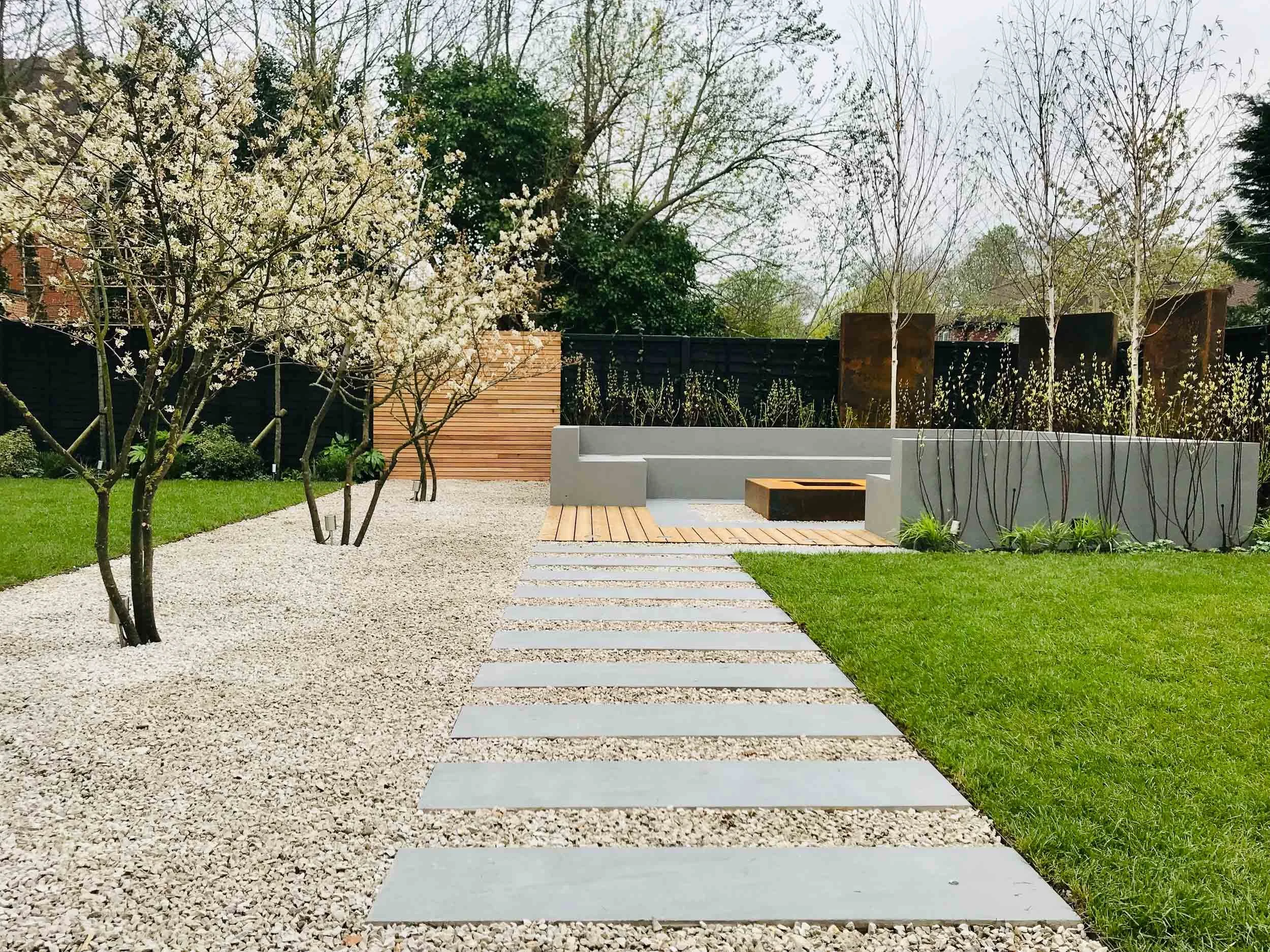 Modern backyard with a gravel walkway, wooden deck, green grass, blossoming trees, and a seating area with concrete and wooden elements.