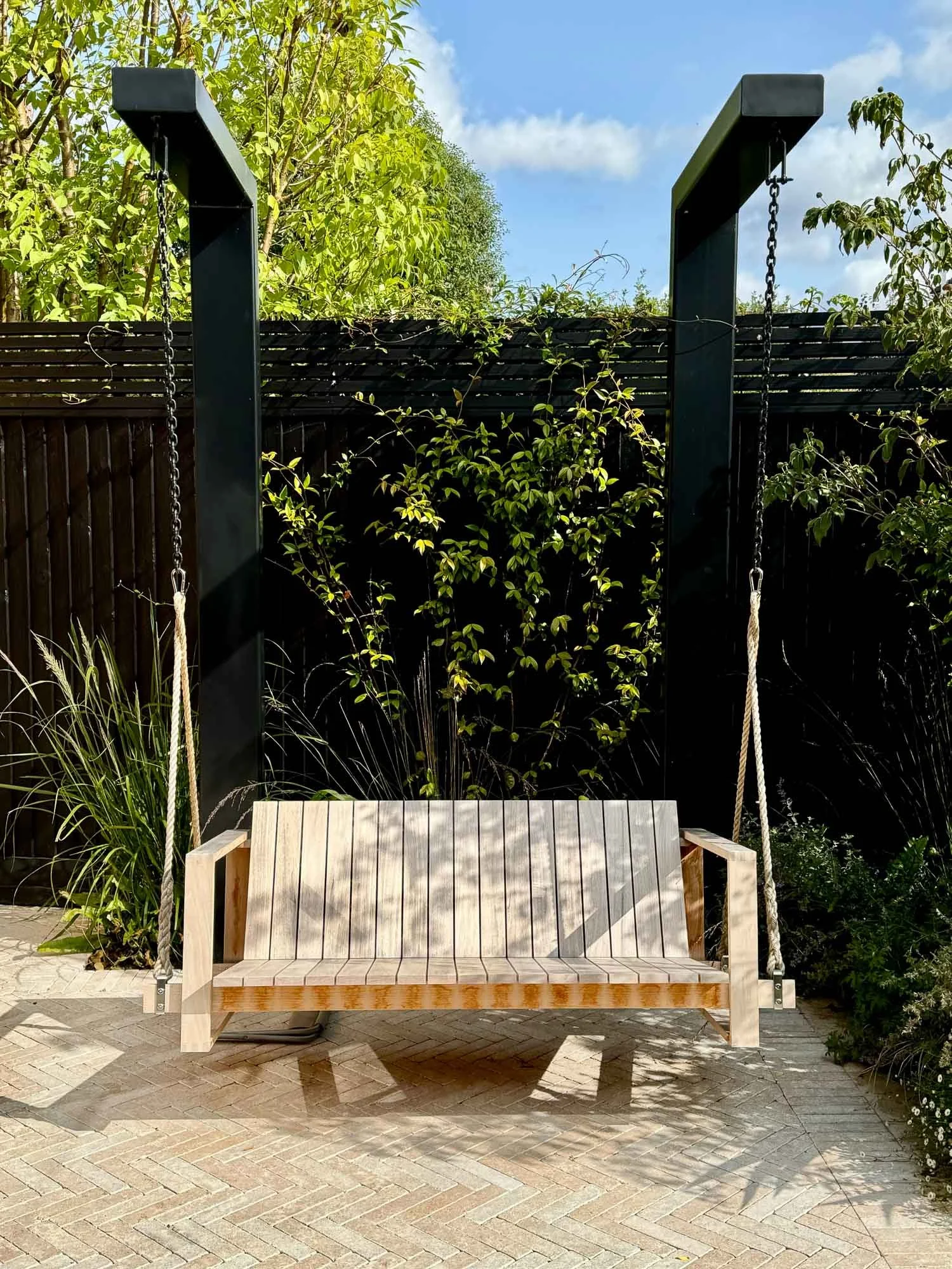 A wooden outdoor swing with beige ropes suspended from a black frame, set against a garden background with green foliage and a brick patio.