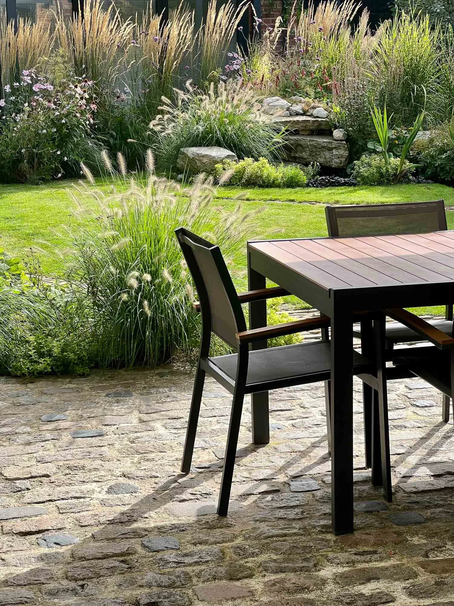 Modern timber dining table and black chairs on a cobbled patio overlooking tall ornamental grasses and pink blooms