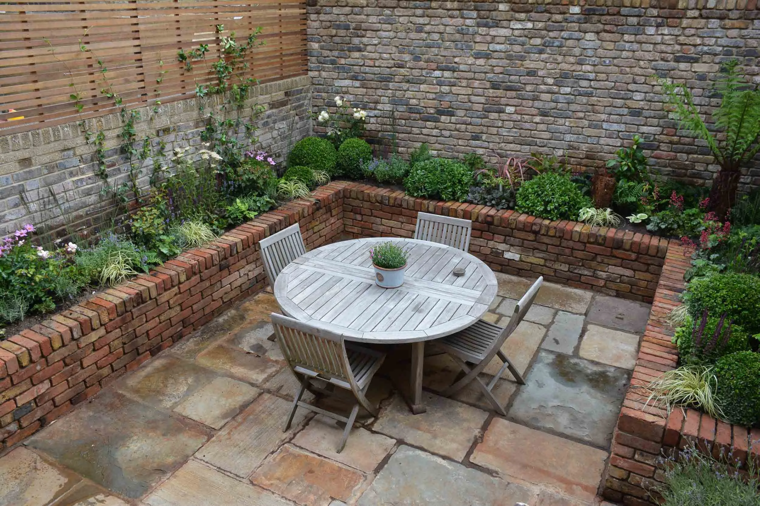 A small outdoor patio with a round wooden table and four matching chairs, surrounded by a brick raised garden bed filled with various green plants and flowers. The patio floor is made of large stone slabs, and the background features wooden and brick