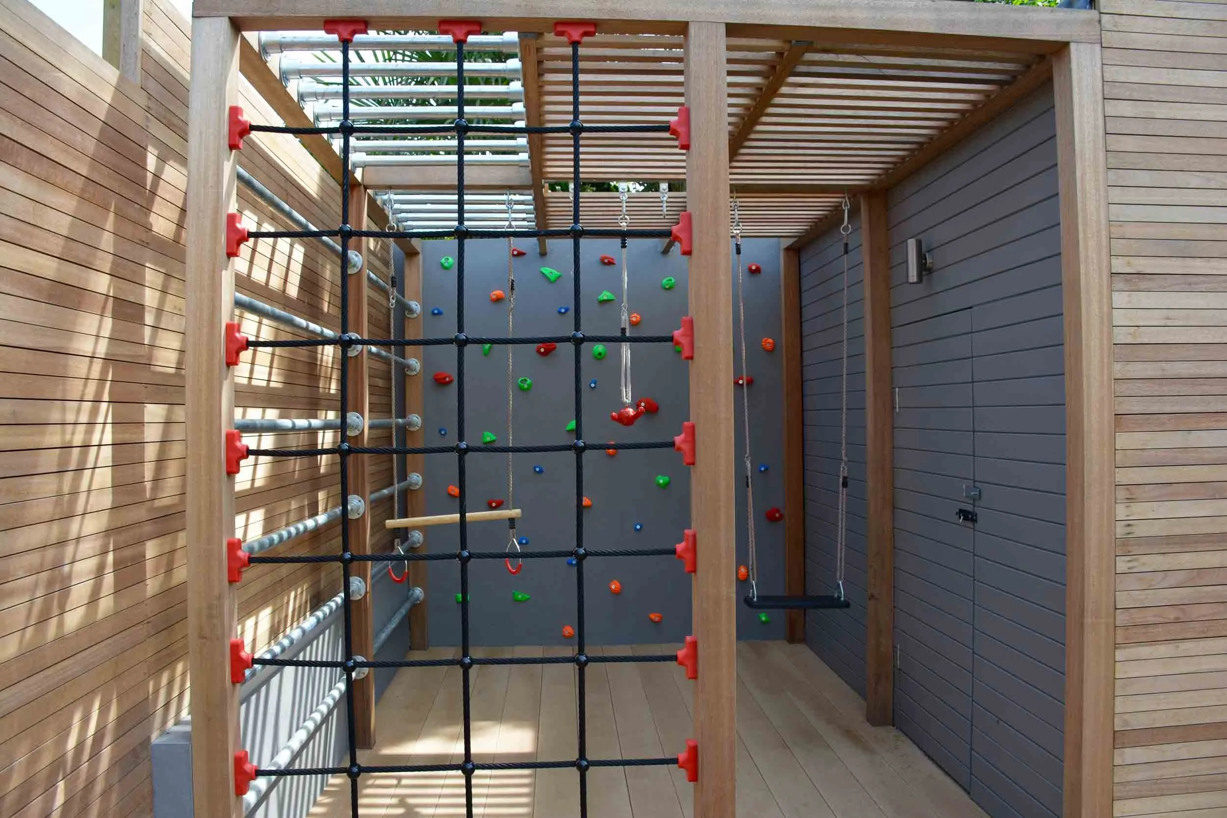 Indoor outdoor play area with climbing wall, swing, and wooden structures with sliding surfaces and safety netting.