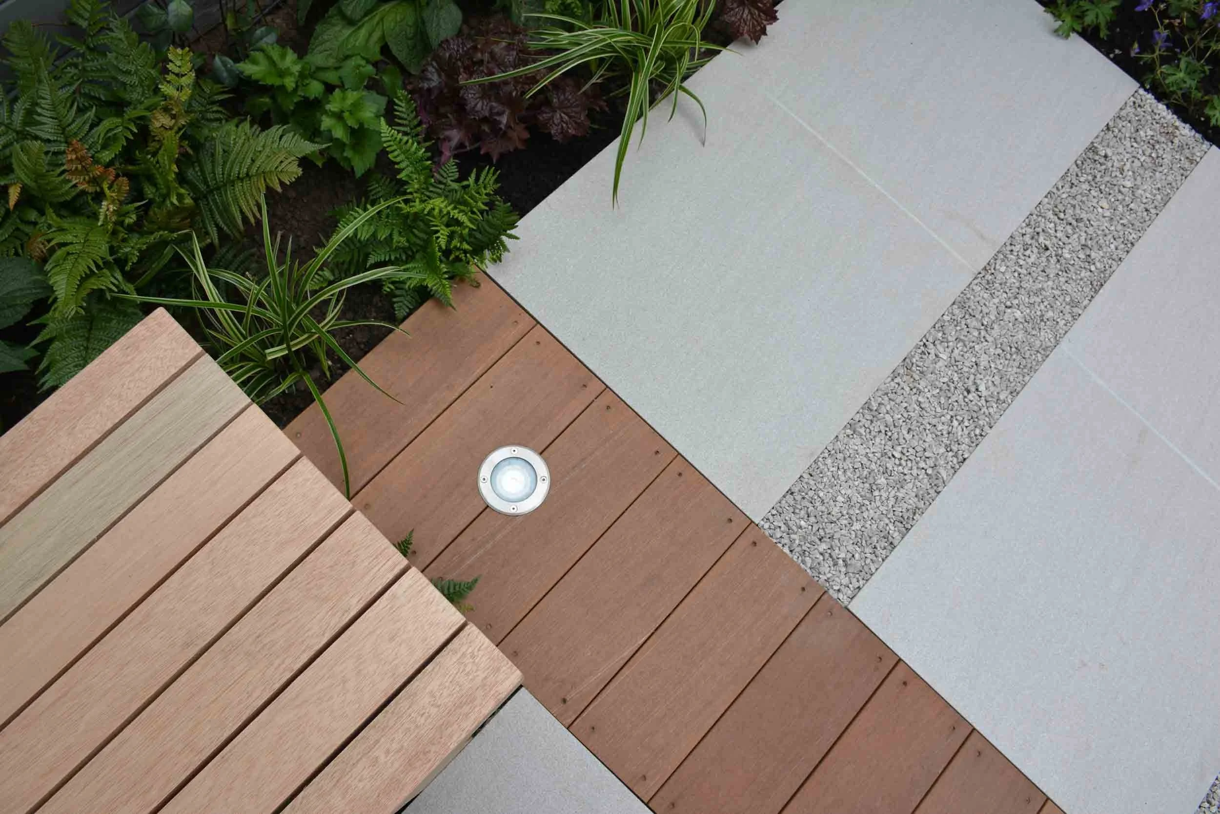 A section of outdoor flooring with a mix of wooden planks, concrete tiles, and gravel, bordered by lush green plants.