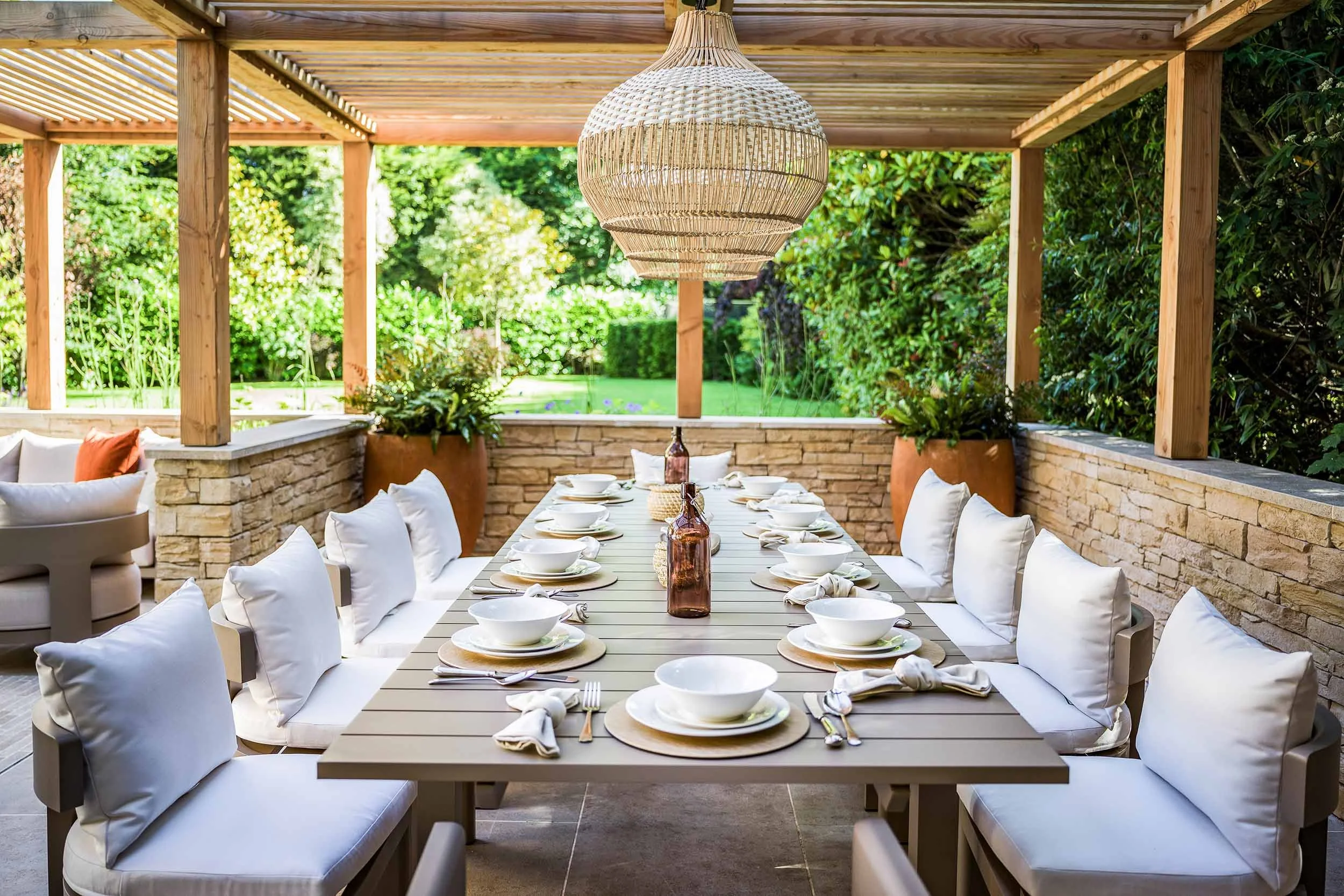 Outdoor dining area with a long table set for a meal, surrounded by white cushioned chairs, under a wooden pergola. The table has bowls, plates, napkins, and bottles, with large potted plants and greenery in the background.