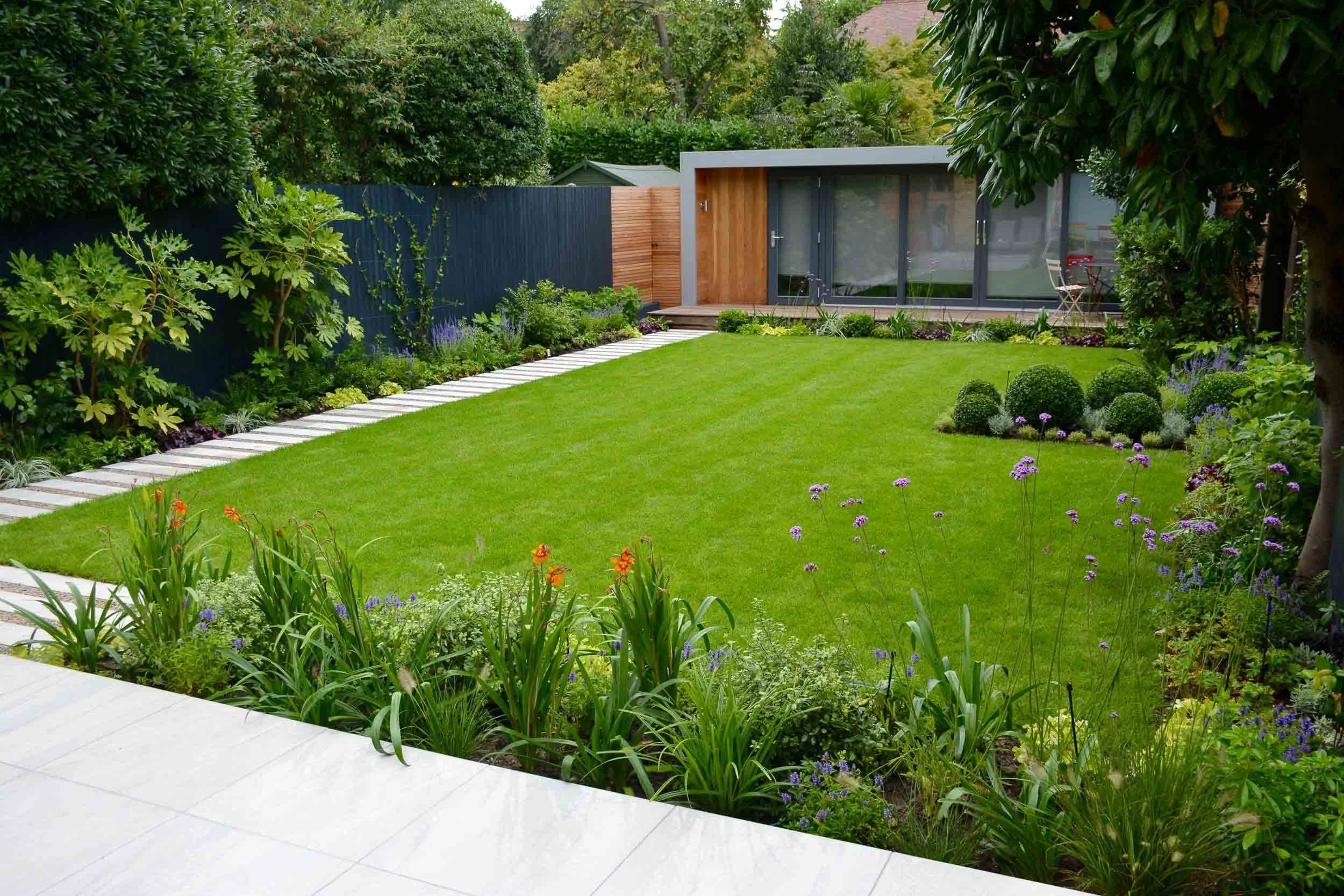 A well-maintained backyard with a lush green lawn, surrounded by a variety of plants, shrubs, and trees. There's a modern building with large glass doors and a patio area in the background, bordered by a dark fence. A pathway made of rectangular whit