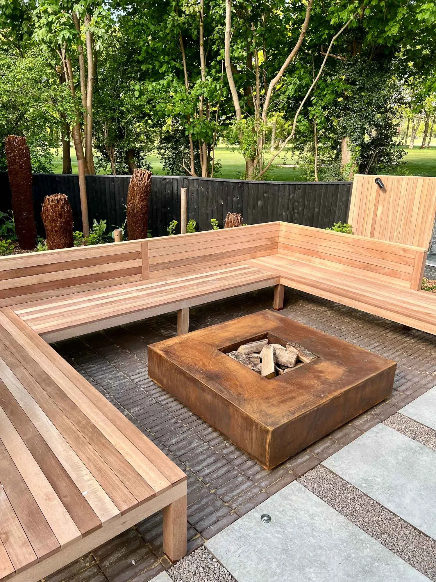 An outdoor seating area with a built-in wooden bench surrounding a fire pit, with trees and a wooden fence in the background.