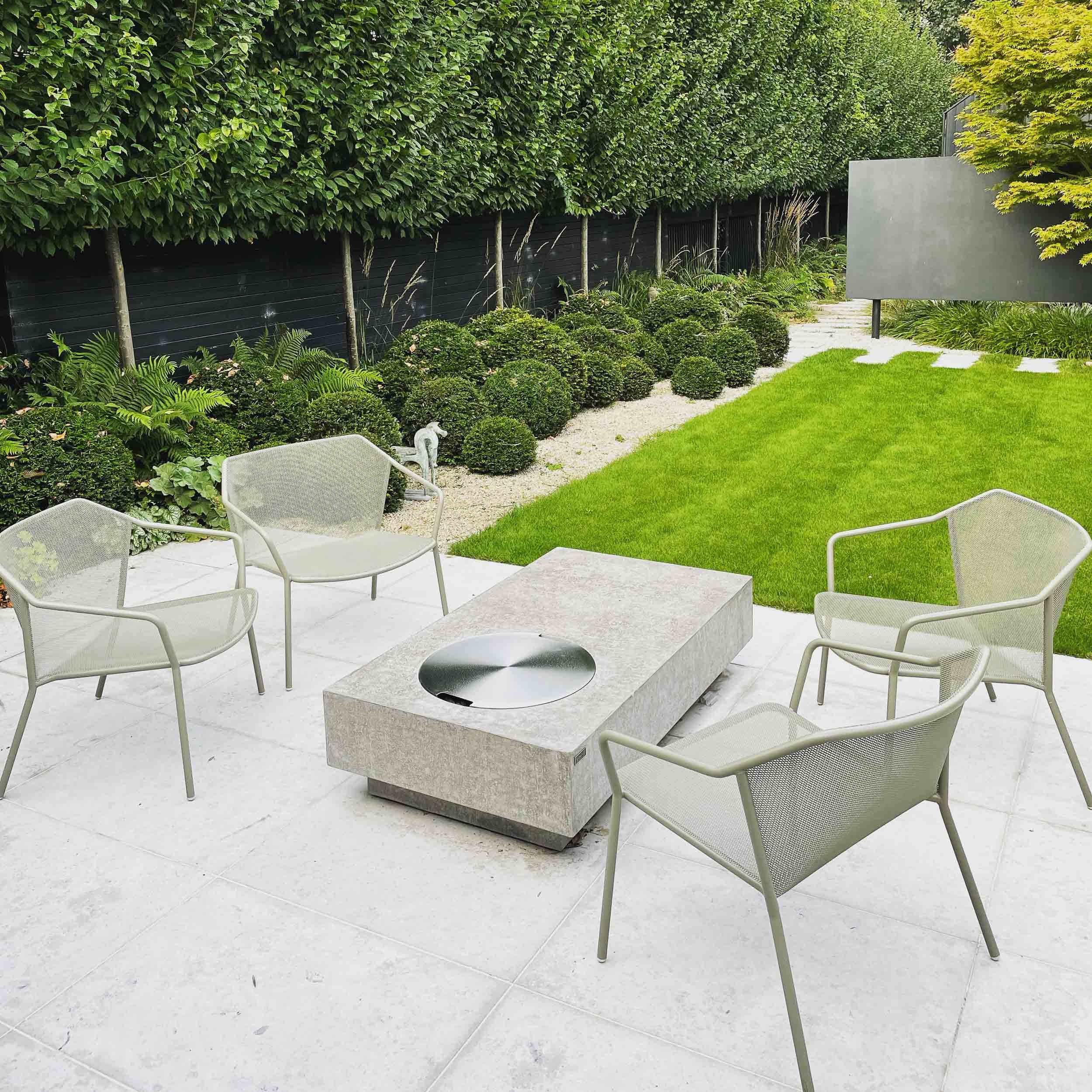 Modern outdoor garden with a rectangular fire pit, four beige mesh chairs, manicured grass, and lush greenery.