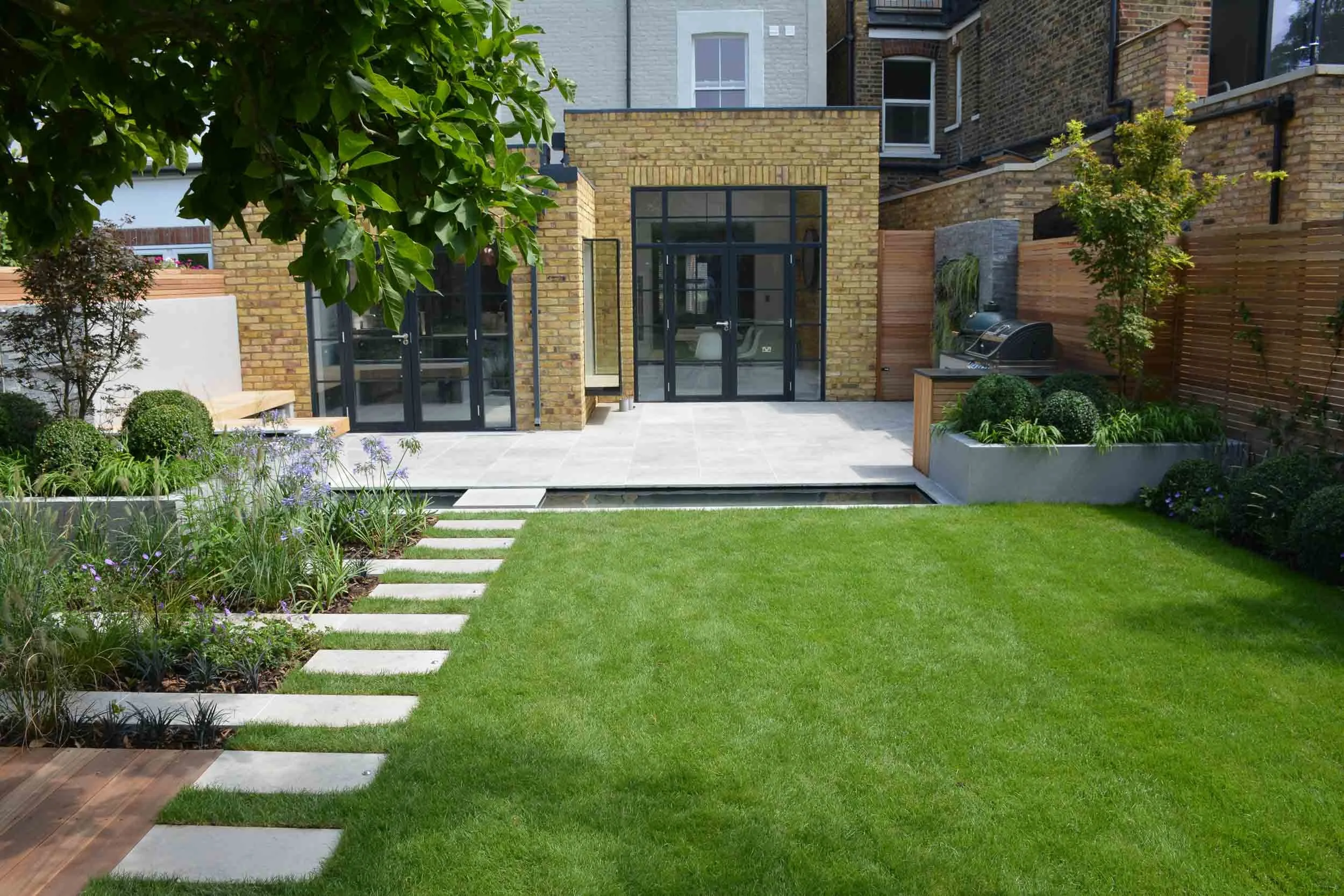 Backyard with green grass, stepping stones, landscaped flower beds with plants, and a patio with glass doors and windows leading into a brick building.