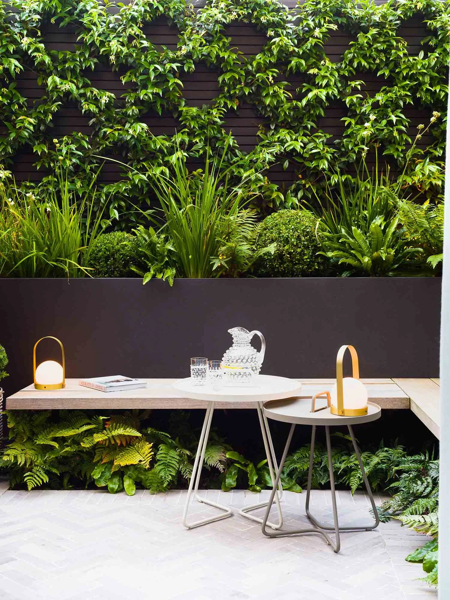 Intimate seating area with a small table, two lanterns on a patterned patio, backed by a lush ivy wall and planter