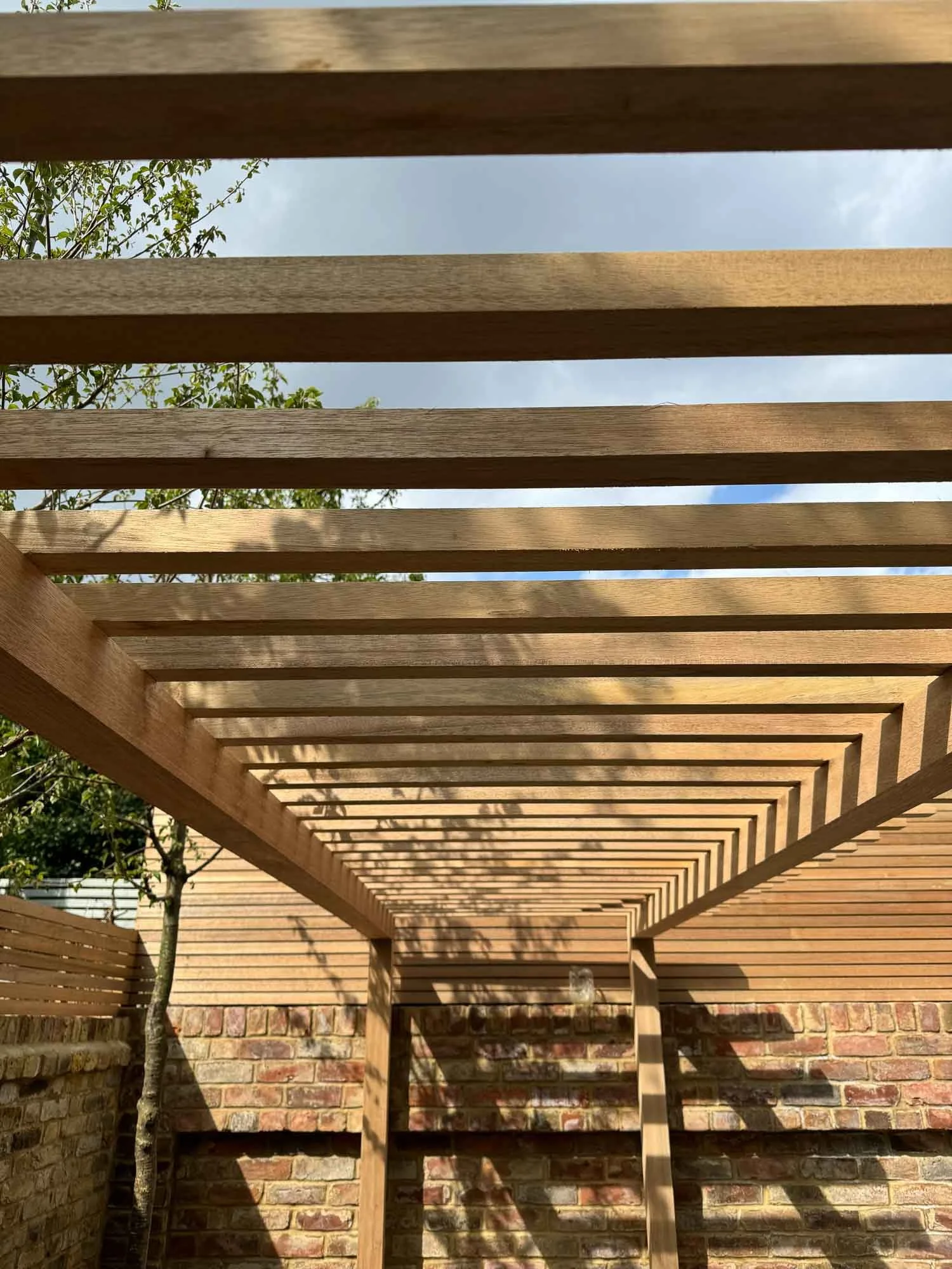 A wooden pergola with evenly spaced horizontal beams casts shadows on a brick wall and a small tree in a backyard.