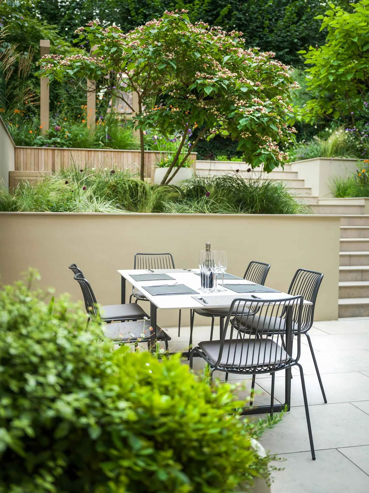 Sunken terrace garden with light grey paving, a wooden L-shaped outdoor sofa, and a wide stone staircase leading to an upper lawn