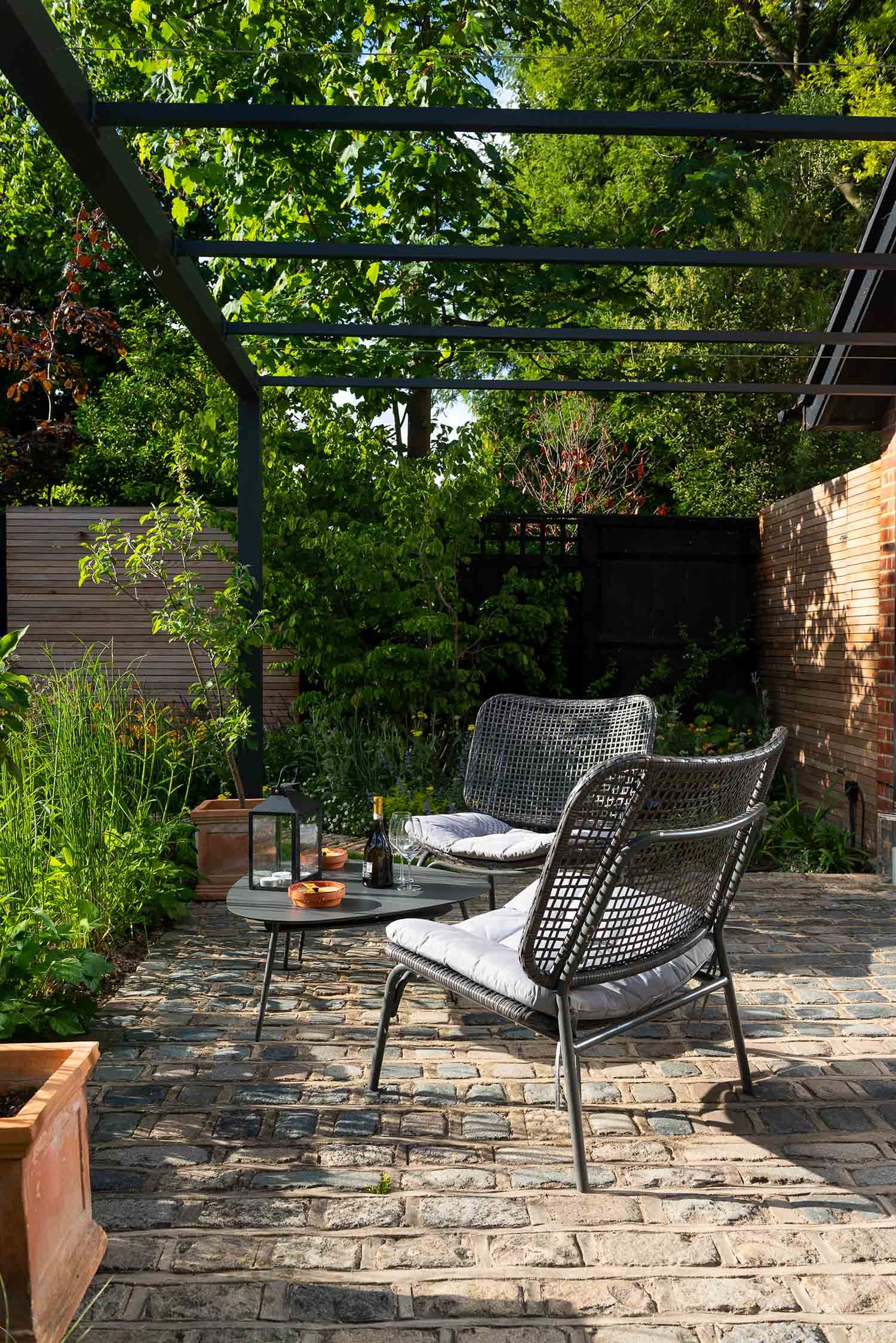 Modern grey wicker chairs and a black metal table on a reclaimed cobbled patio under a contemporary black pergola