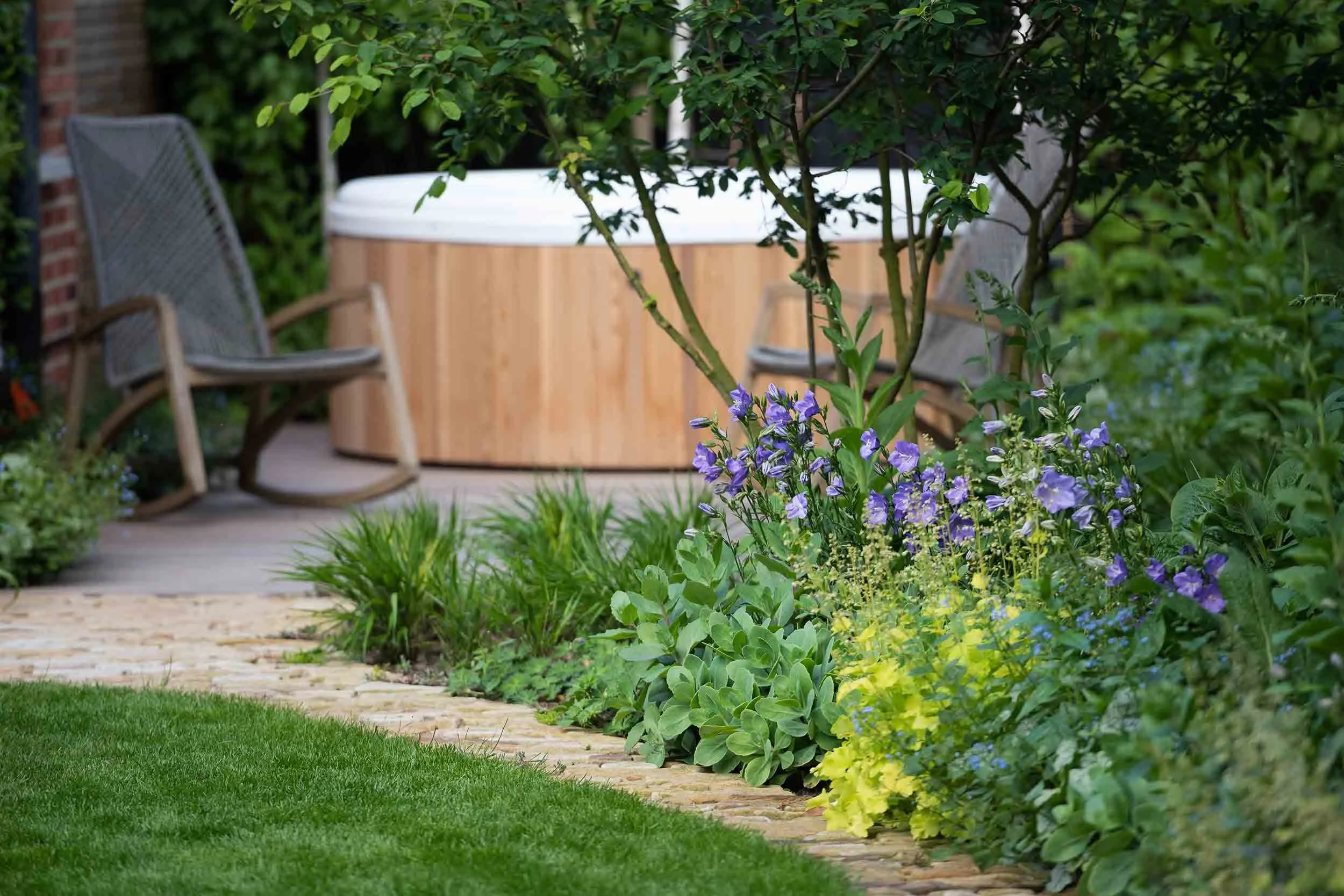 Wooden hot tub and rocking chairs in a secluded garden corner behind a lush border of purple and lime green plants