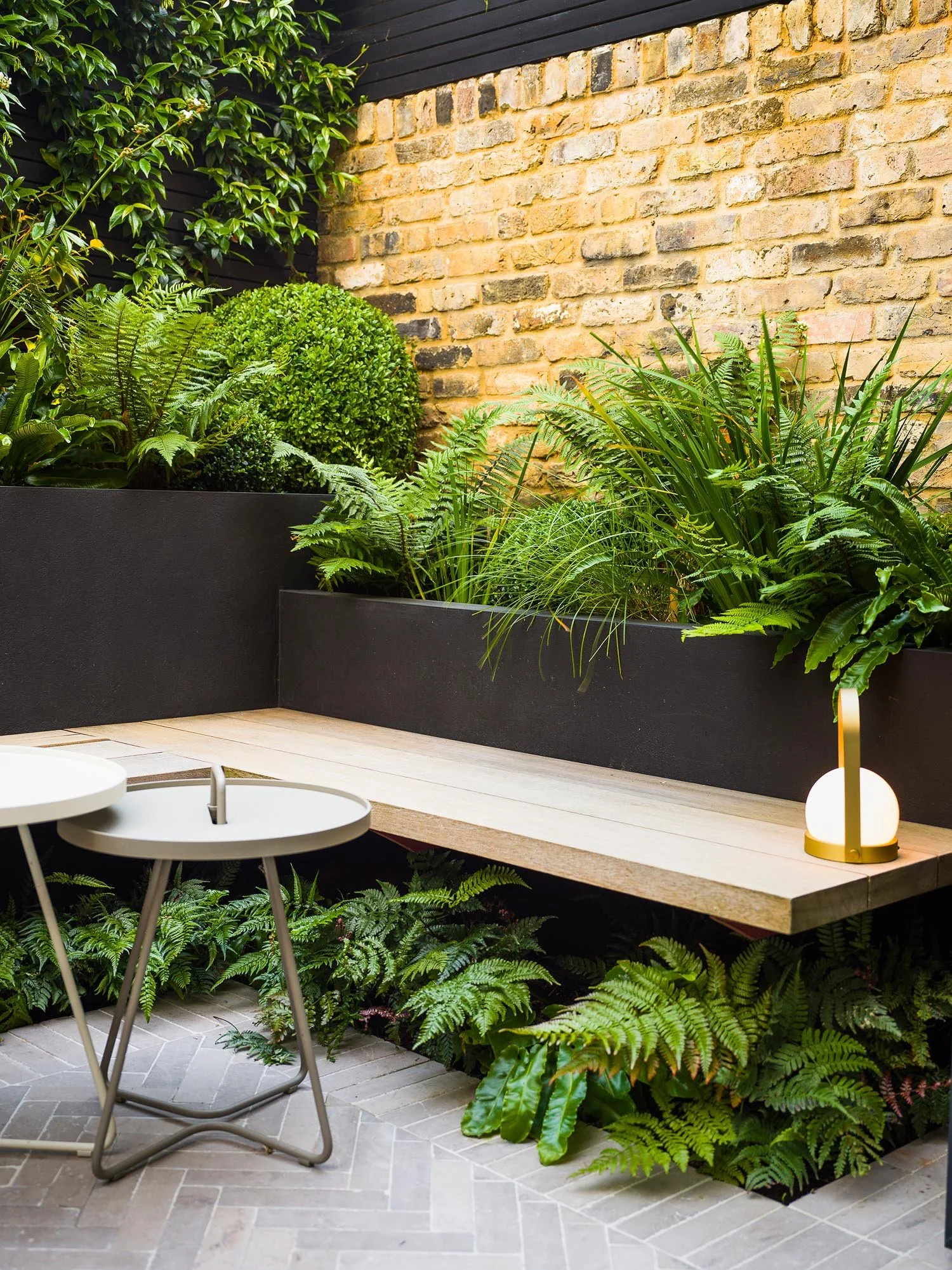Wooden garden bench nestled between a dark grey planter and a brick wall with lush ferns and a glowing orb lantern