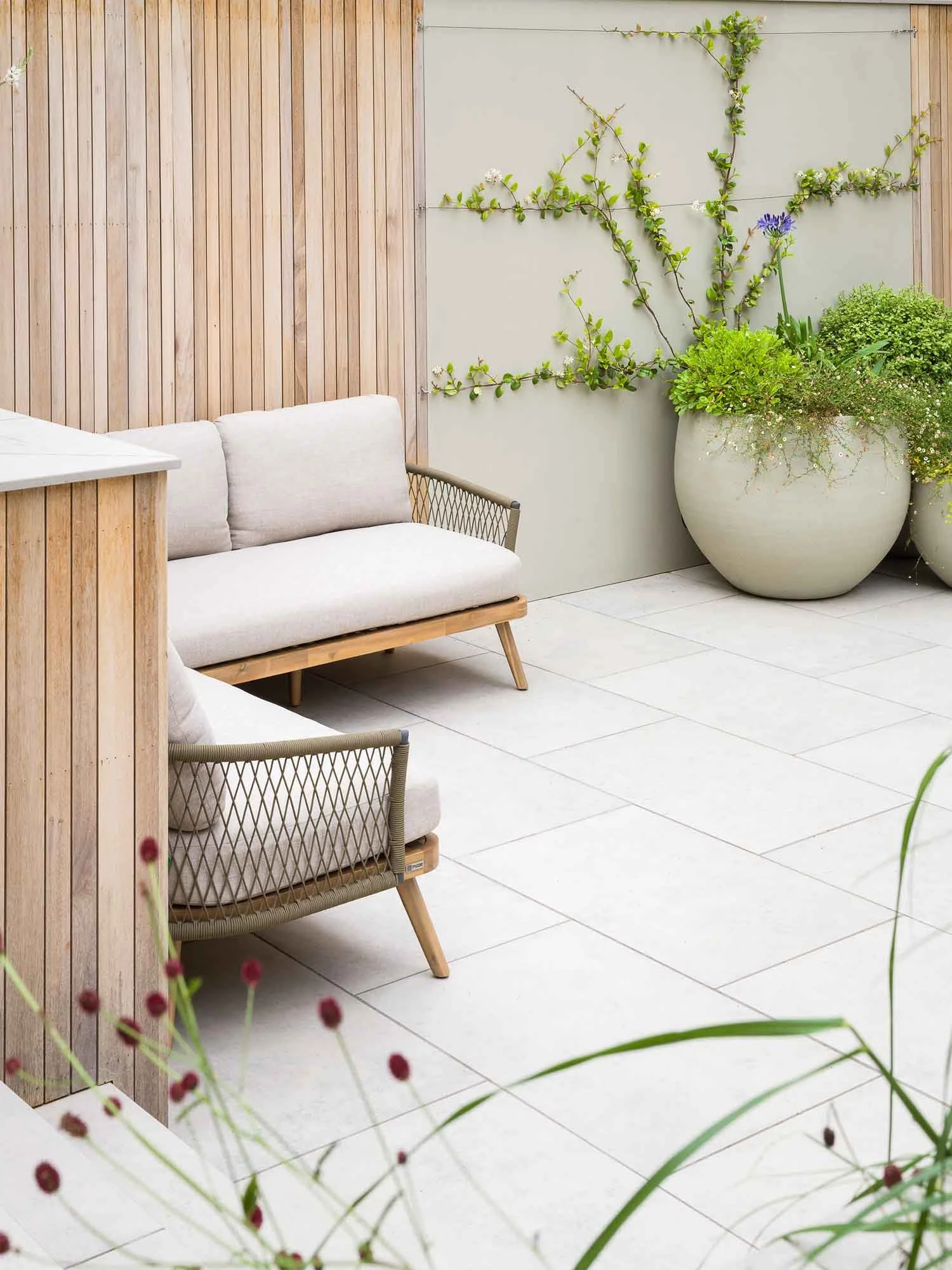 Contemporary outdoor seating area with a wooden sofa on a stone patio with large planters in the background