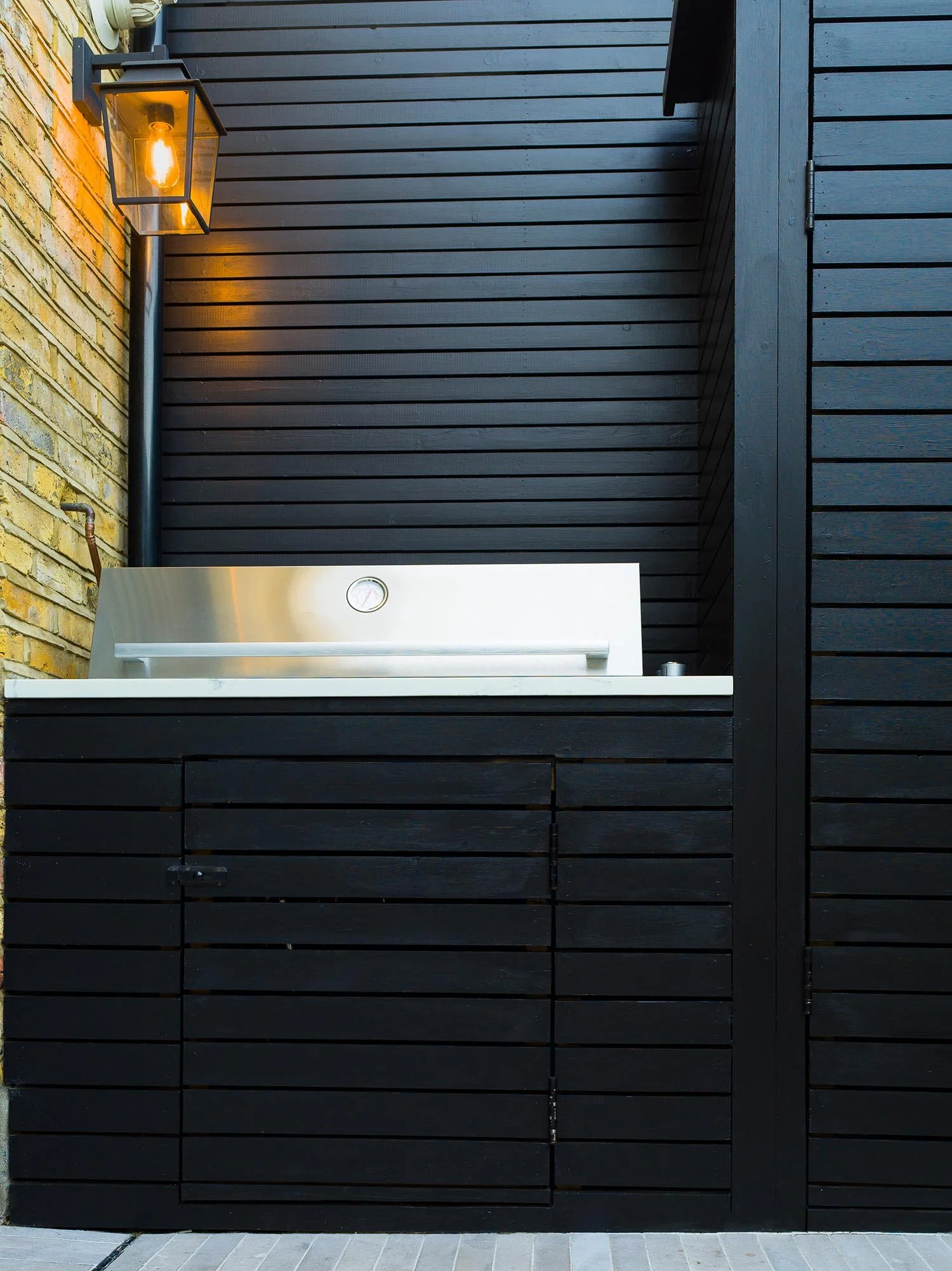 Built in stainless steel gas barbecue integrated into black timber cabinetry with an atmospheric lit wall lantern