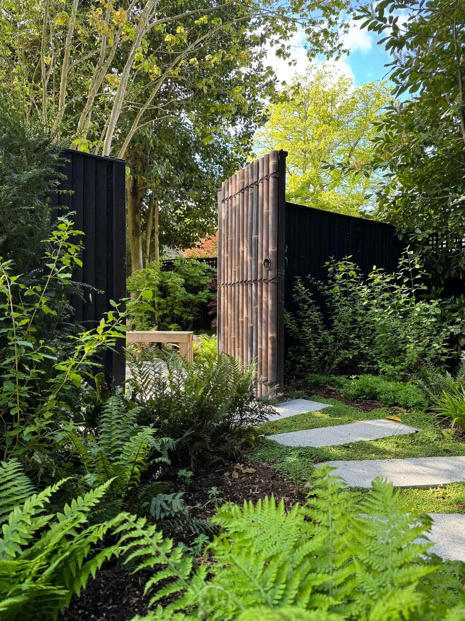 A garden with lush green plants and ferns, a pathway made of stone slabs, and a wooden gate opening to a larger garden area with trees and blue sky.