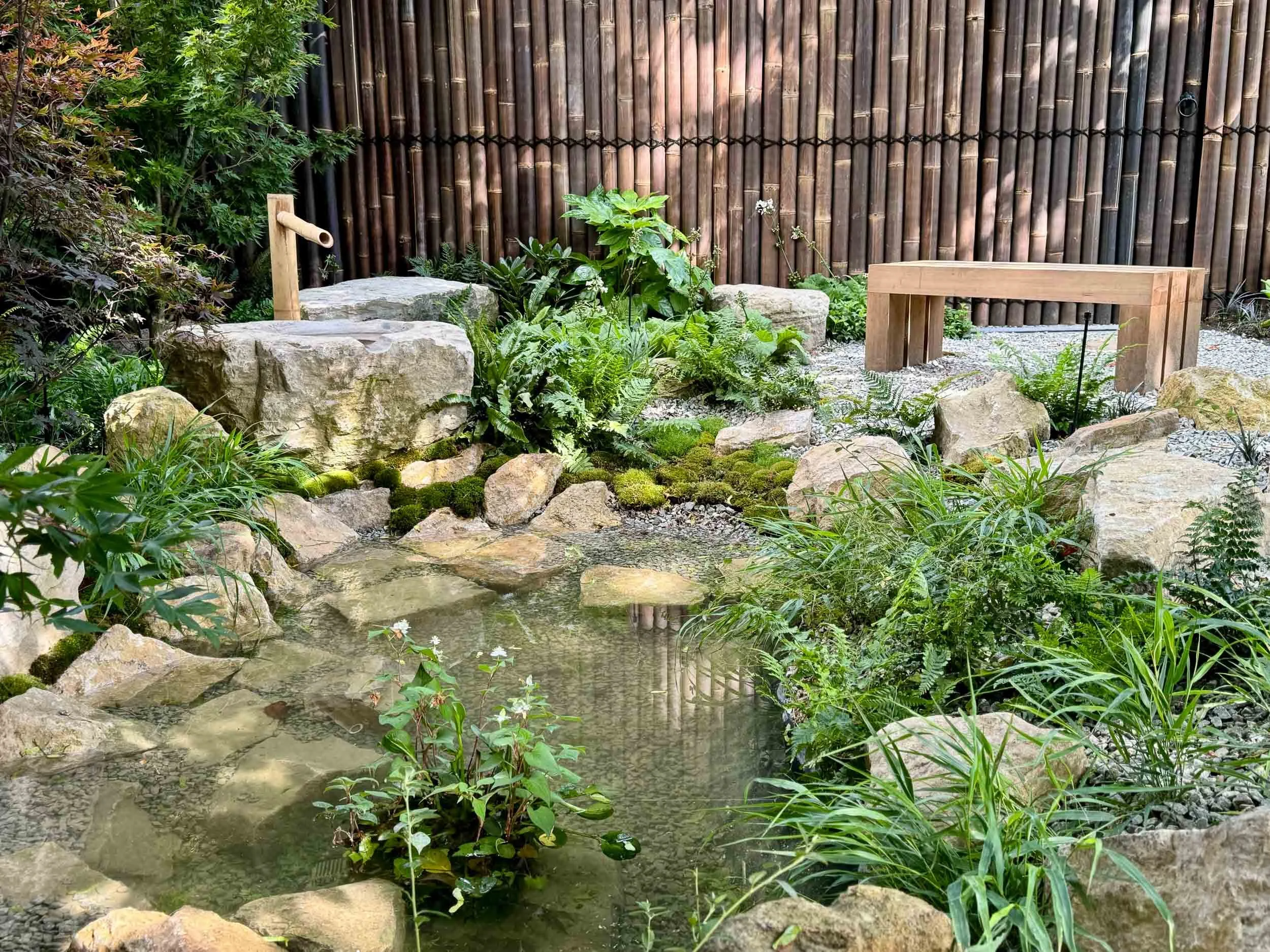 A small landscaped garden with a pond, large rocks, lush green plants, and a wooden bench and water spigot in the background, enclosed by a bamboo fence.