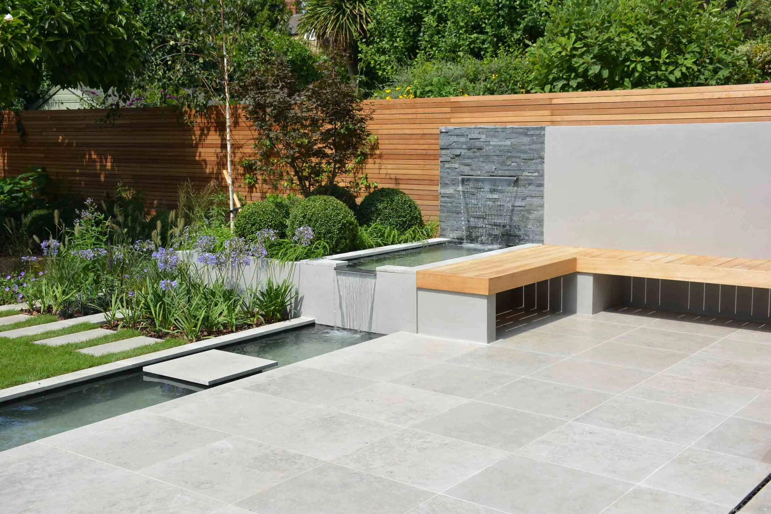 Modern backyard with a water feature, stone waterfall, wooden bench, tiled patio, and lush green plants and shrubs, enclosed by a wooden fence.