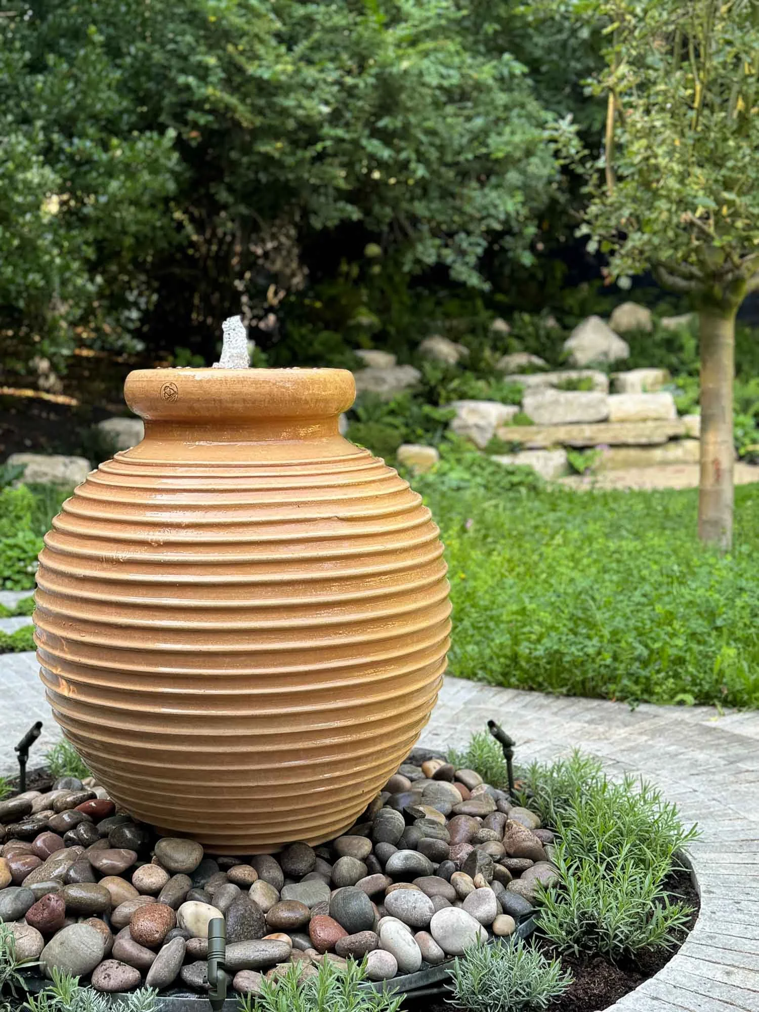 A large tan ceramic vase with horizontal ridges sits on a bed of smooth, multicolored rocks in a garden with lush greenery and trees in the background.