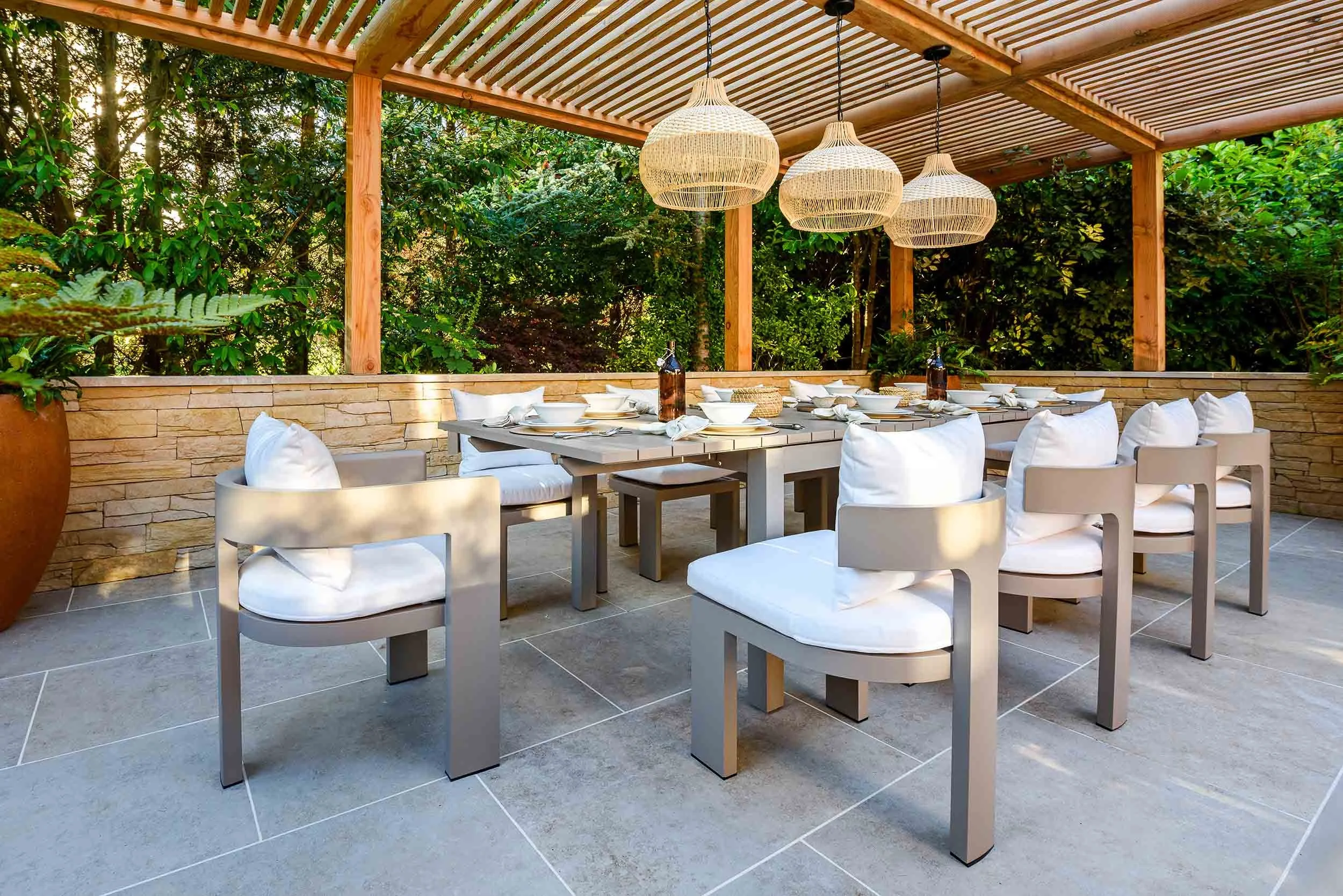 Outdoor dining table with white chairs and woven pendant lights under a timber pergola on a grey stone patio