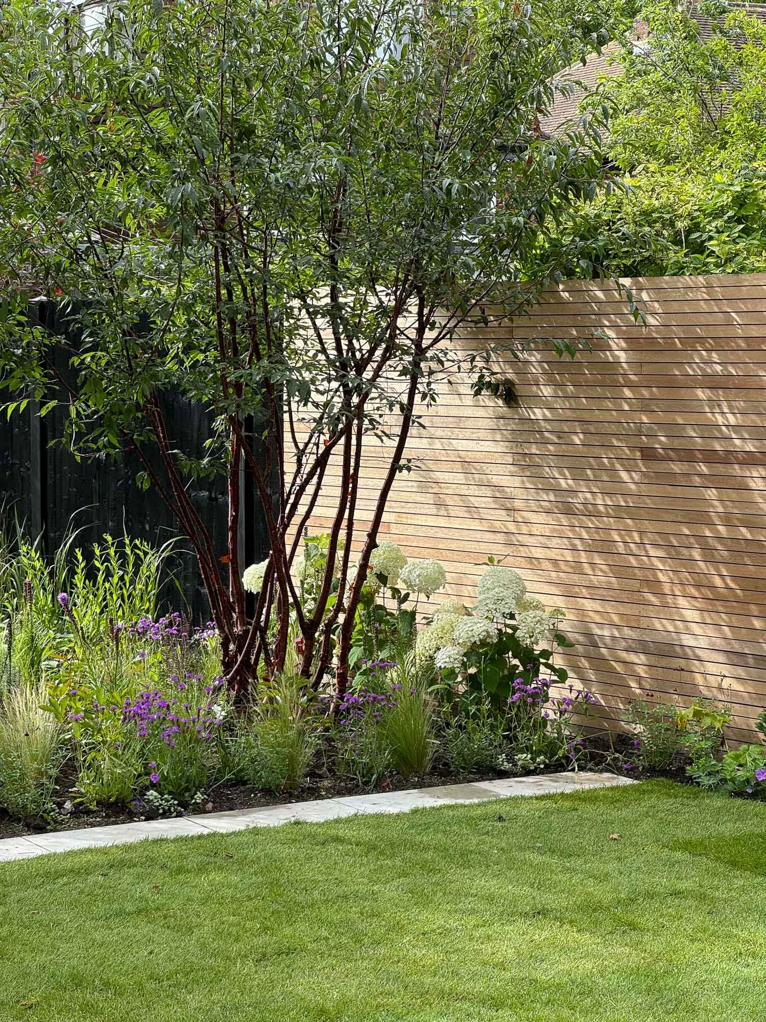 Backyard garden with a tree, purple and white flowers, green grass, a wooden fence, and a beige wall.