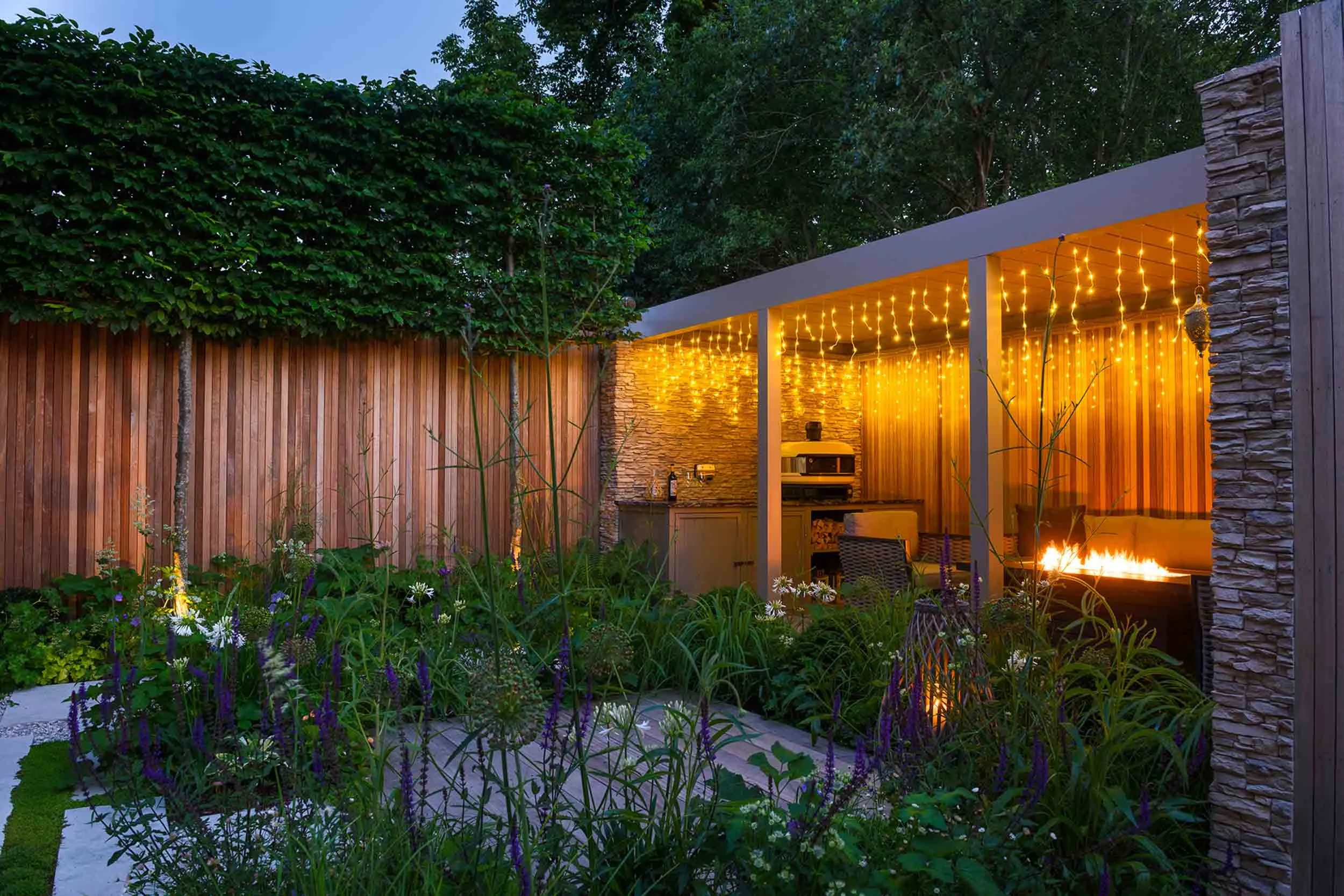 Evening view of the garden pergola and seating area, illuminated by warm yellow string lights