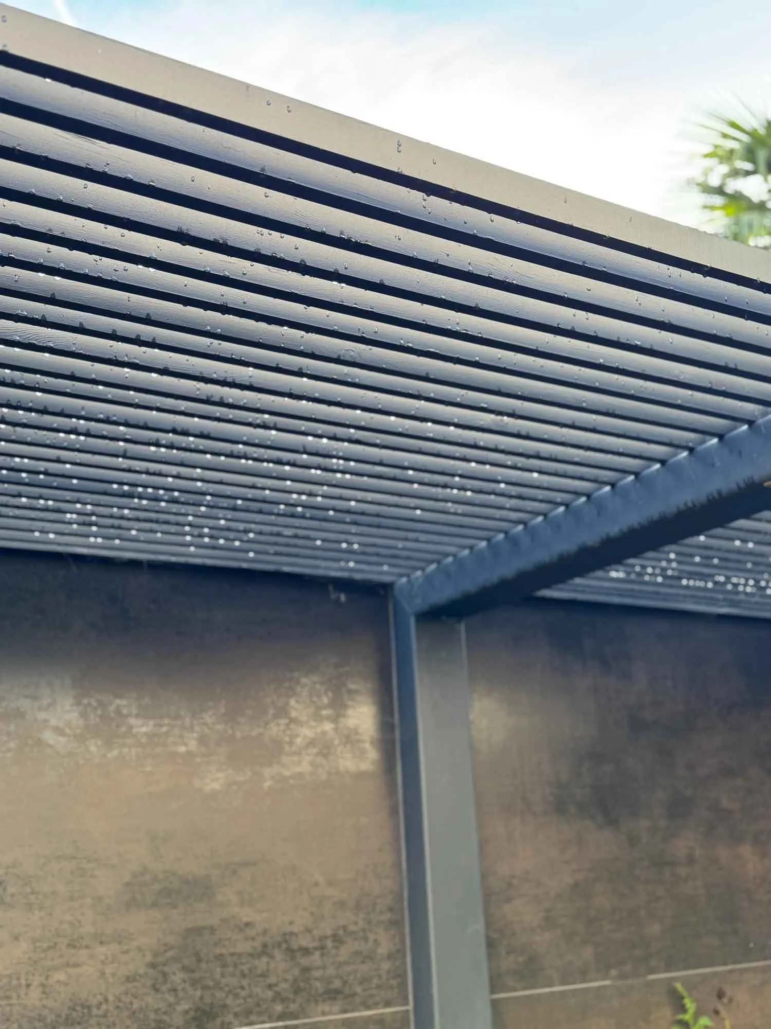 Close-up of a metal structure with horizontal slats, drizzling with rain, and a background of a blue sky and greenery.