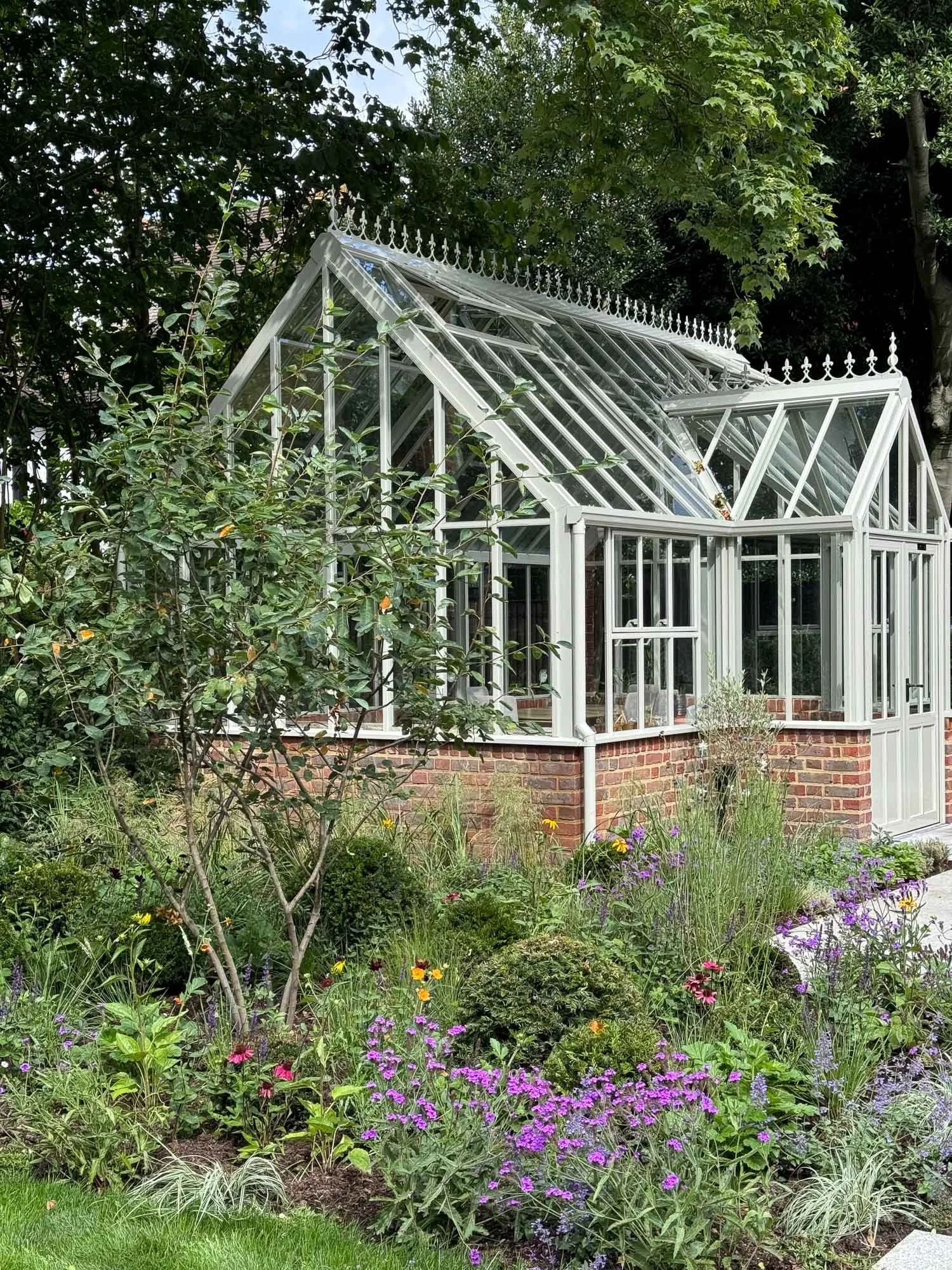 A greenhouse with glass panels, white frame, and decorative trim, surrounded by a lush garden with various flowering plants and trees.