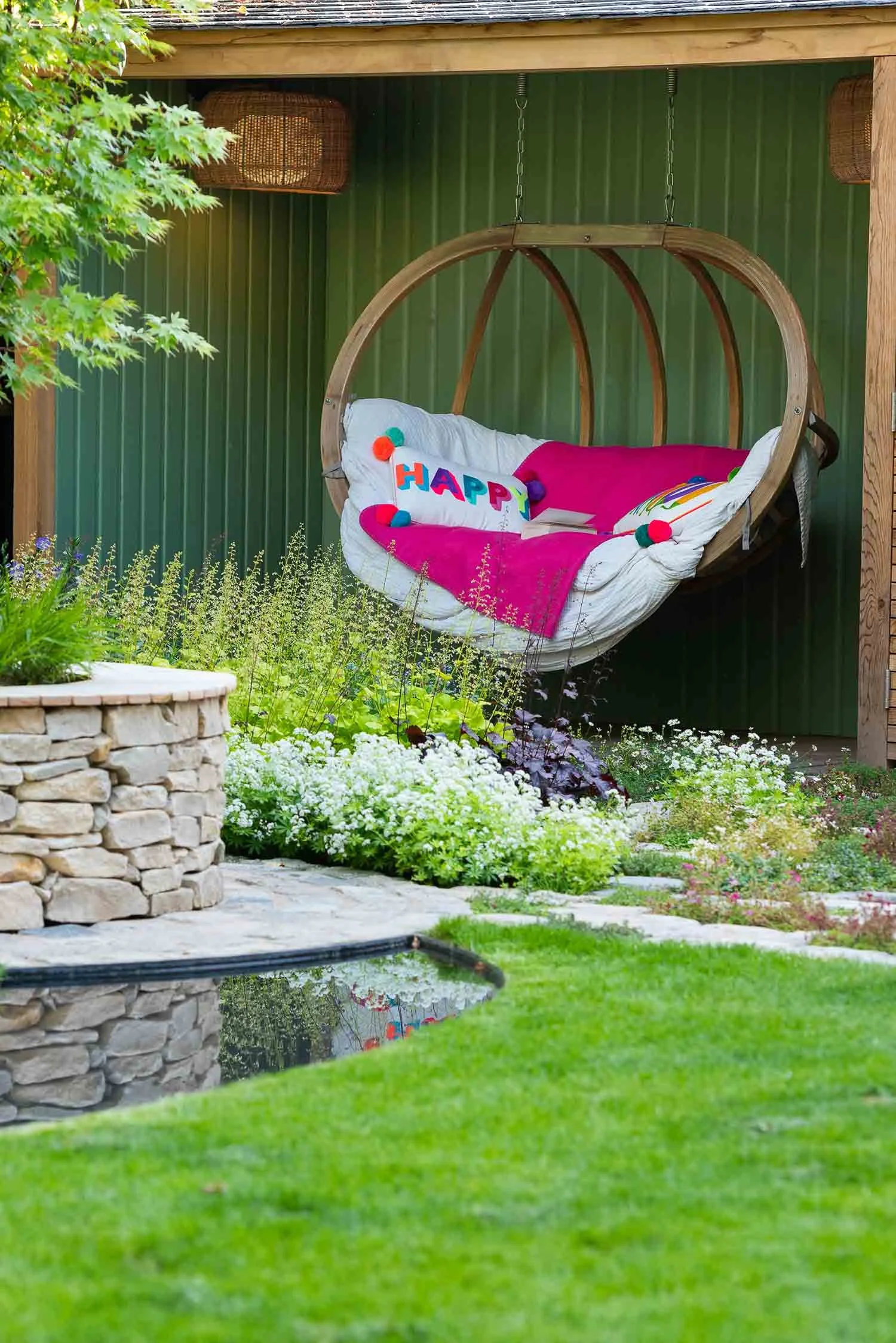 Hanging wooden garden chair with pink blankets and white pillows under a green timber porch near a stone wall