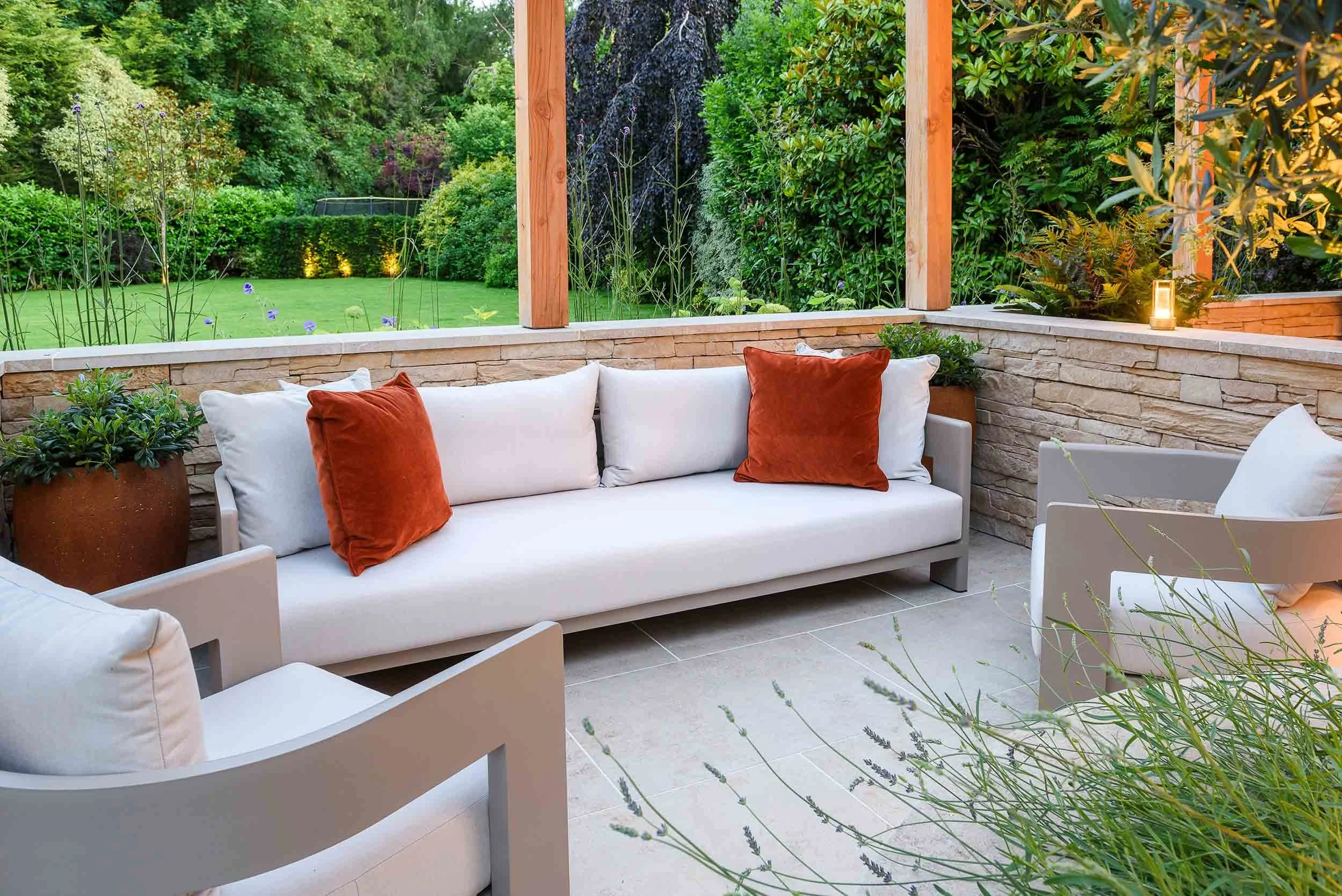 Outdoor lounge area with a grey sofa and orange cushions on a stone patio enclosed by low dry stone walls