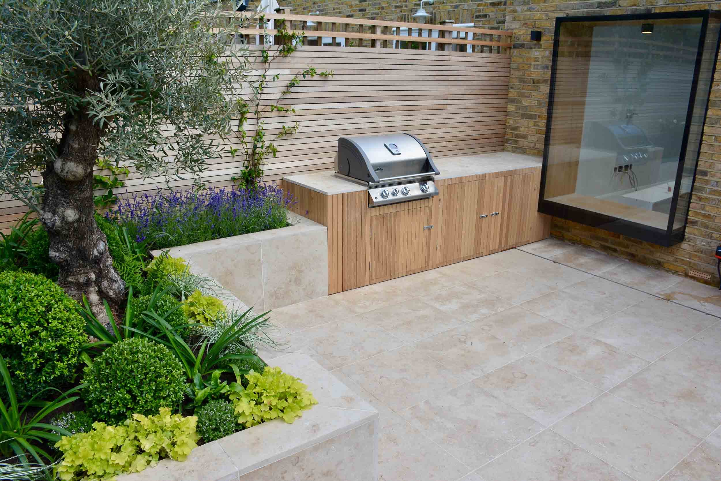 Outdoor patio with built-in grill, wooden cabinetry, potted plants, and a garden area with plants and trees.