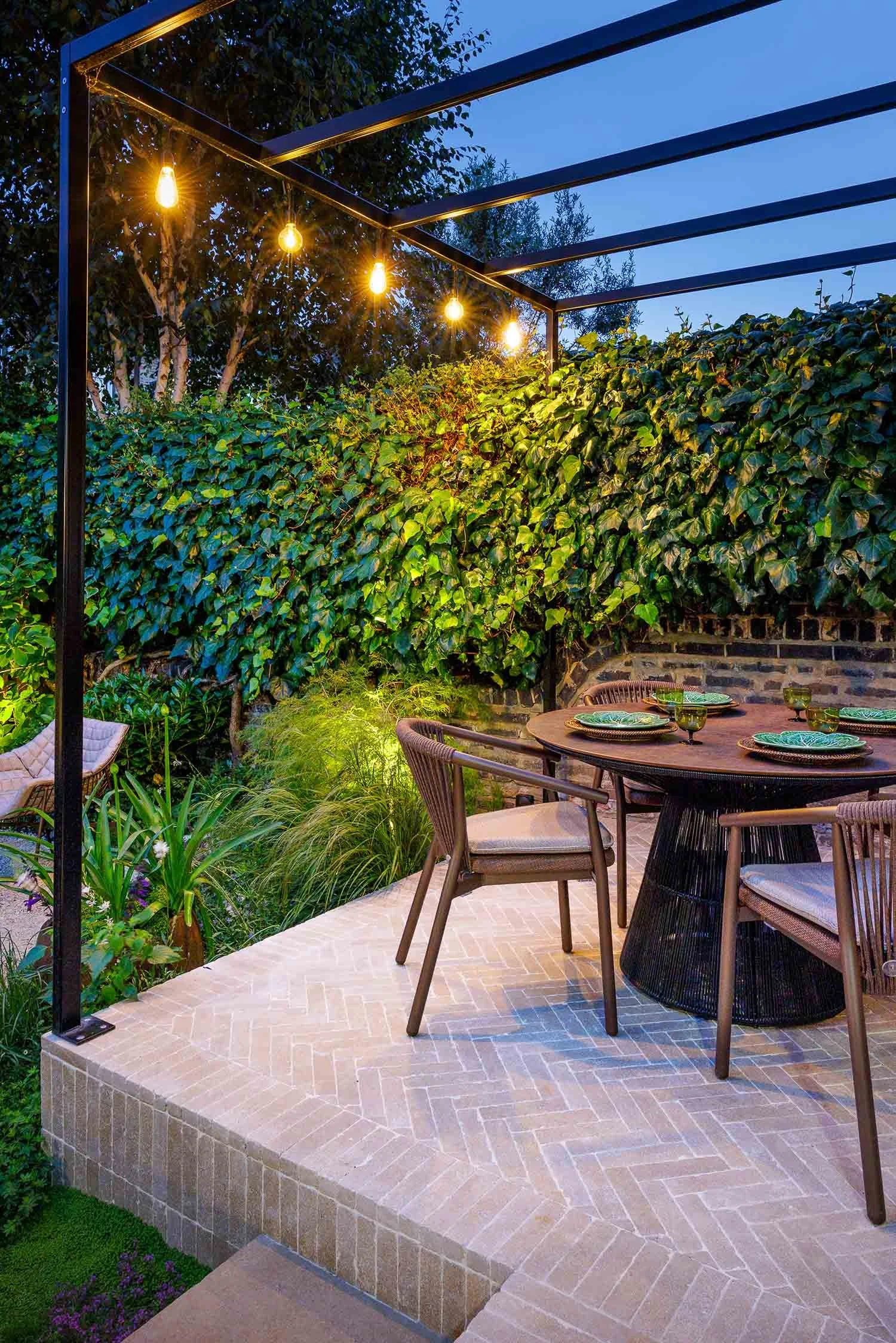 Garden dining table at night under a metal pergola with glowing festoon lights against an ivy wall