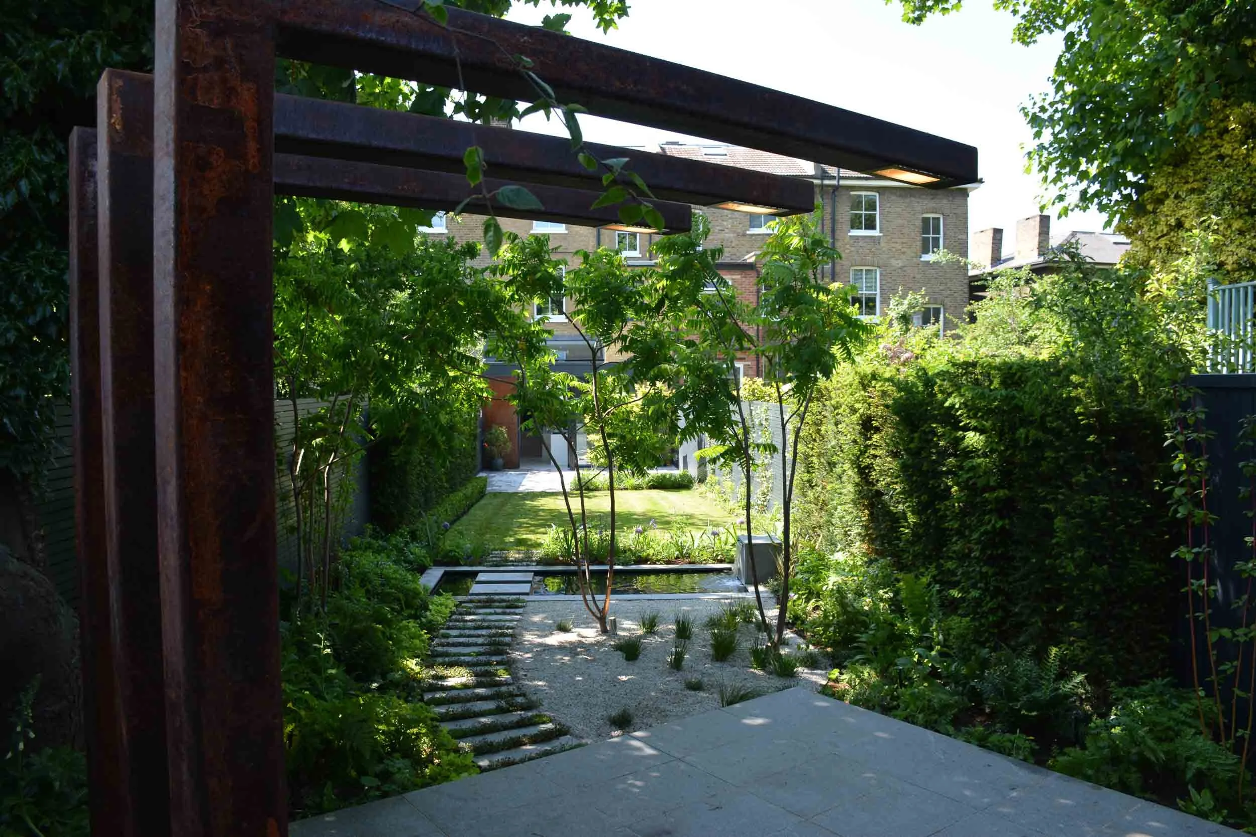 A lush, green urban backyard garden with a narrow stone pathway, trees, shrubs, and a modern residential building in the background.