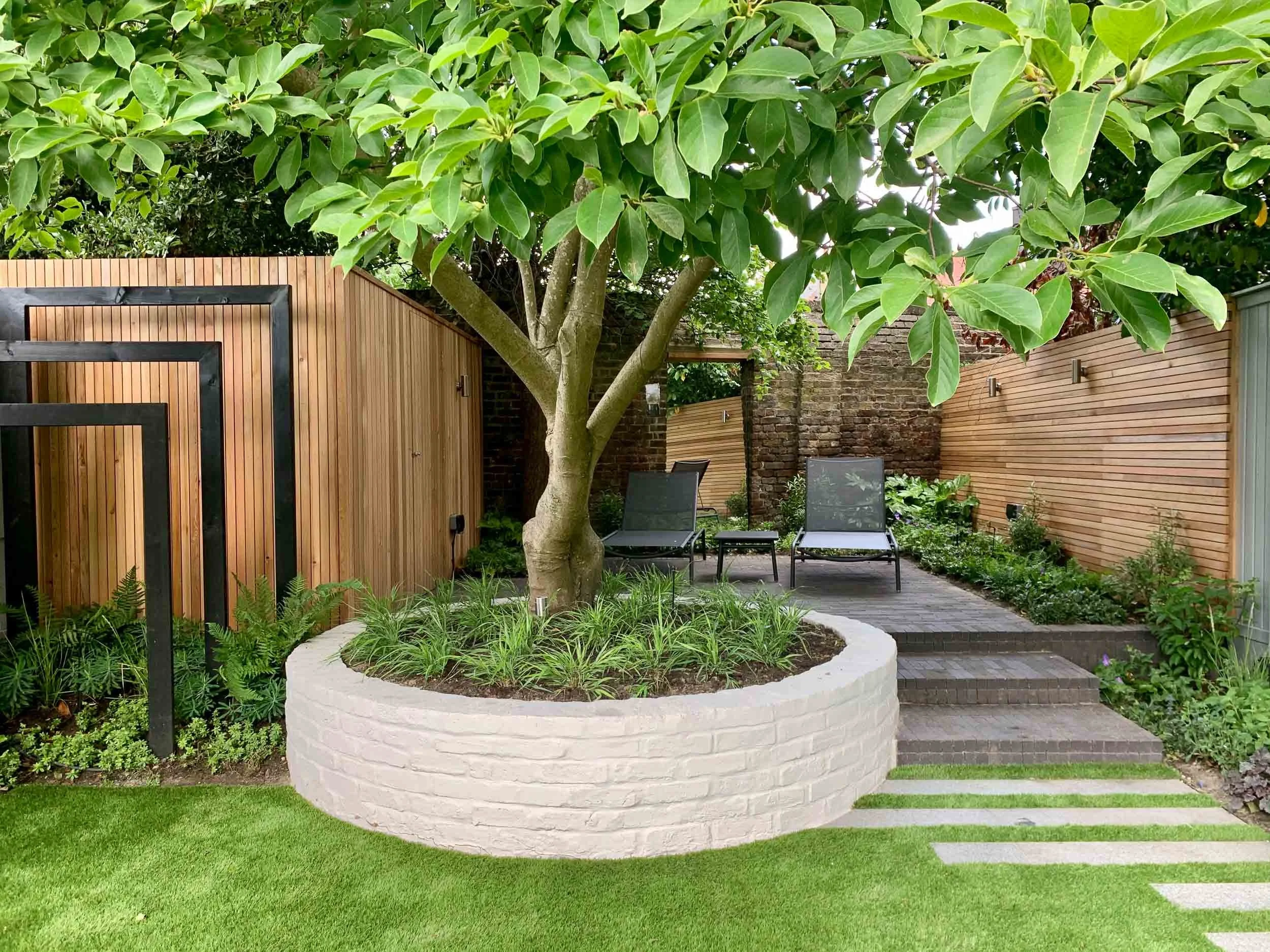 A modern backyard garden with a large tree in a white circular planter, surrounded by green grass, wooden fencing, a brick pathway, and two lounge chairs.